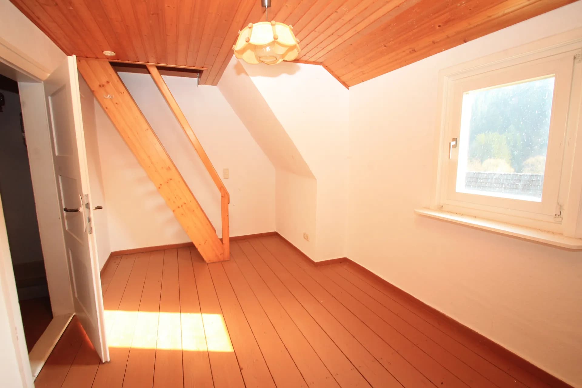 Gemütliches Dachgeschosszimmer mit Holzdeckenverkleidung, Dielenboden, Aufgang zum Spitzboden und Tageslichtfenster.