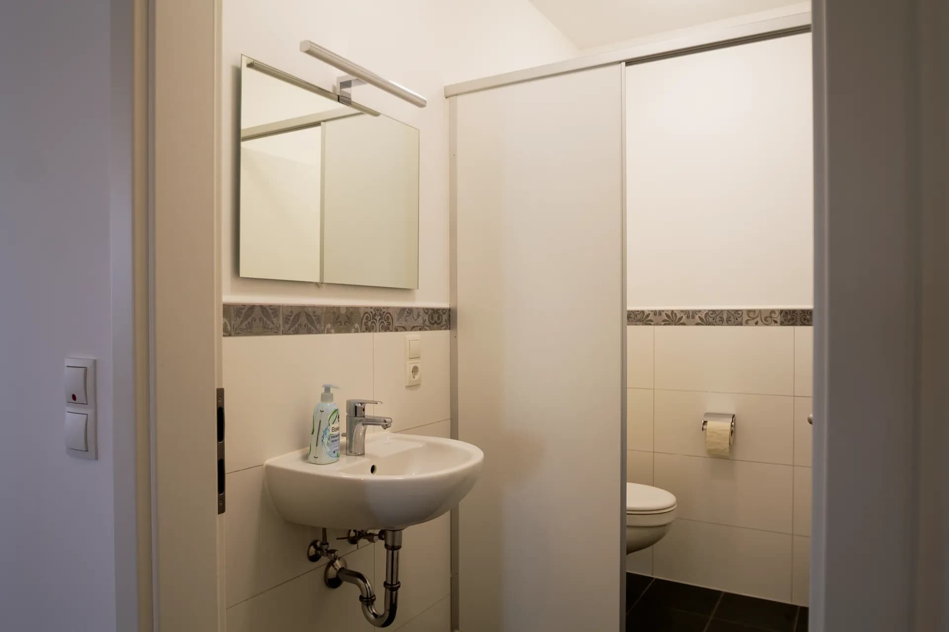 Gepflegtes Büro-WC mit weißen Wandfliesen, Zierbordüre, Handwaschbecken samt Armatur, Spiegelleuchte und abgetrennter Toilettenkabine.
