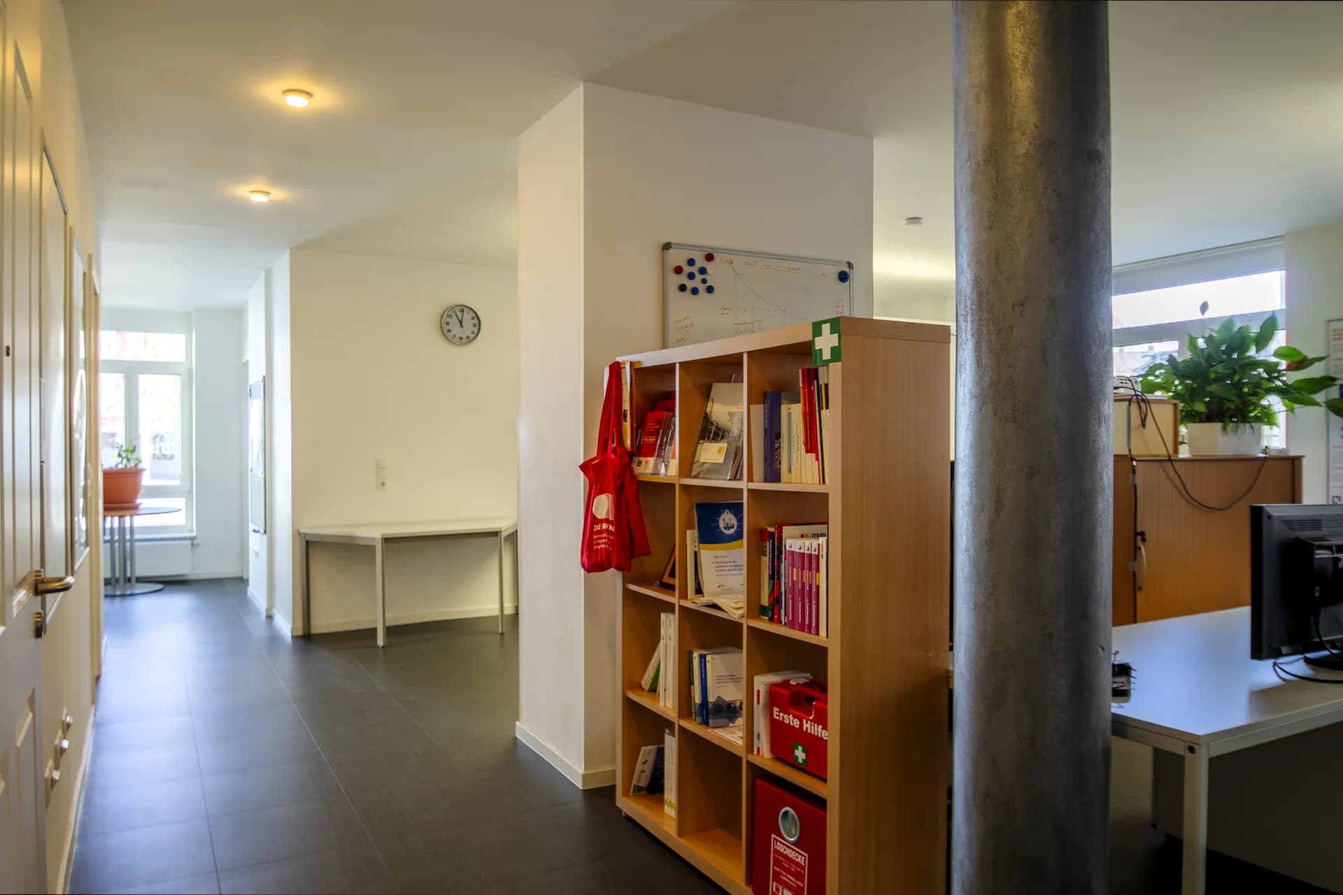 Empfangsbereich und Flur der Büroetage in Hof mit runder Tragsäule, hölzernem Aktenregal samt Erste-Hilfe-Koffer, dunklem Fliesenboden und angrenzenden Arbeitsbereichen.