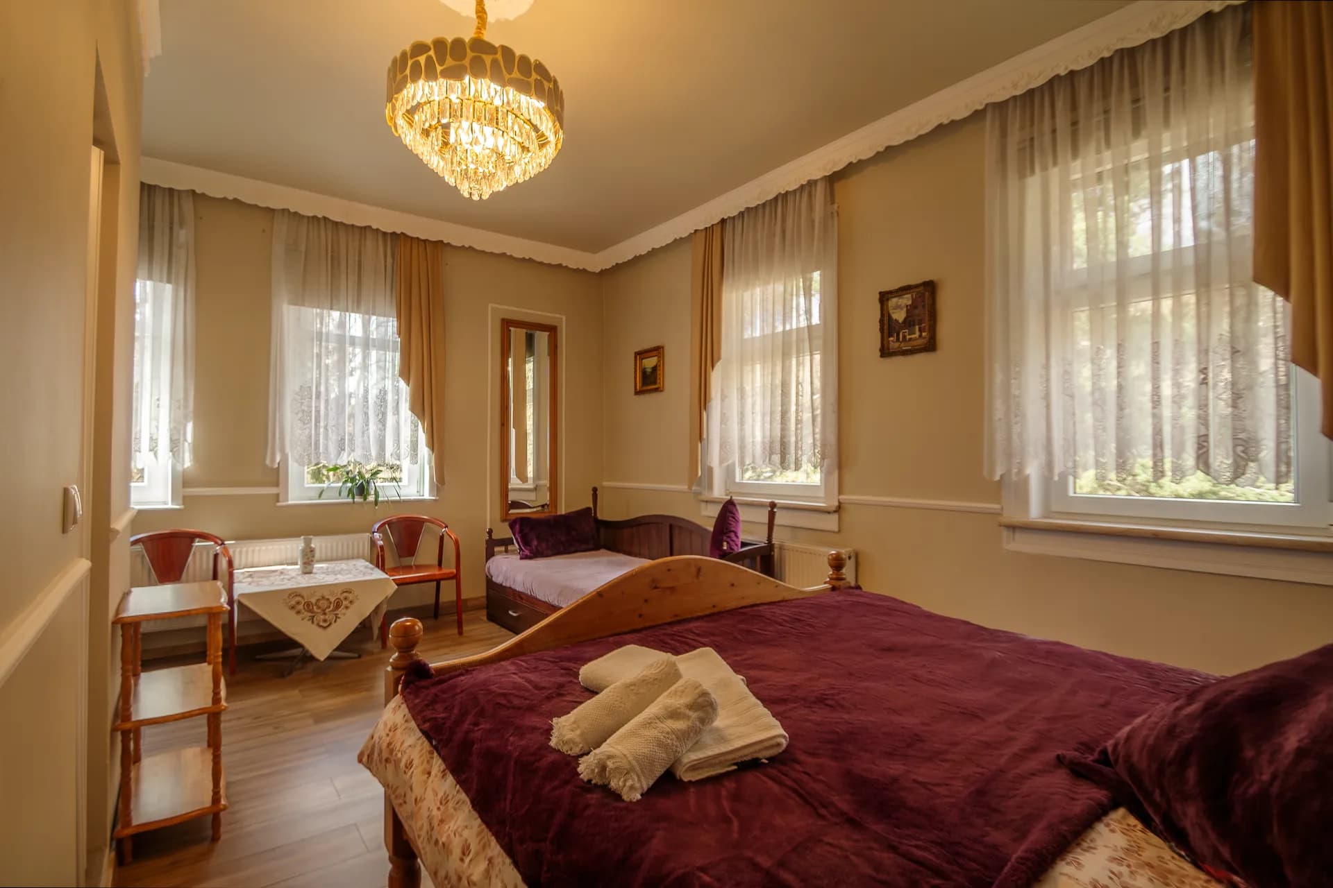 Einladendes Familien- oder Mehrbettzimmer mit Holz-Doppelbett in violetten Tönen, separater Schlafliege, Sitzecke mit Beistelltisch und drei Fenstern mit Ausblick ins Grüne.