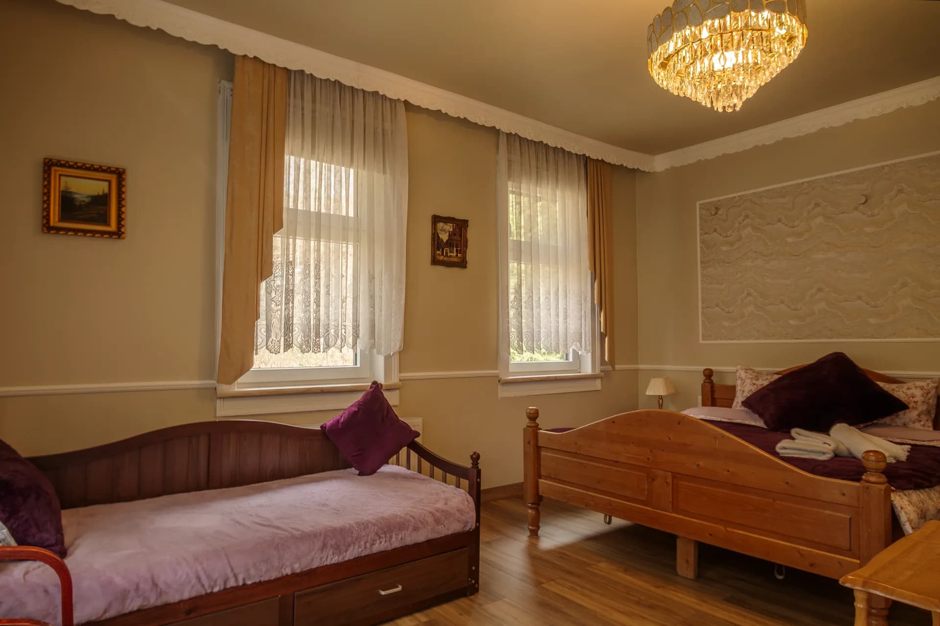 Großzügiges Hotelzimmer mit Laminatboden, Holz-Doppelbett, zusätzlichem Tagesbett/Schlafcouch, funkelndem Kristallkronleuchter und zwei Fenstern mit Vorhängen.
