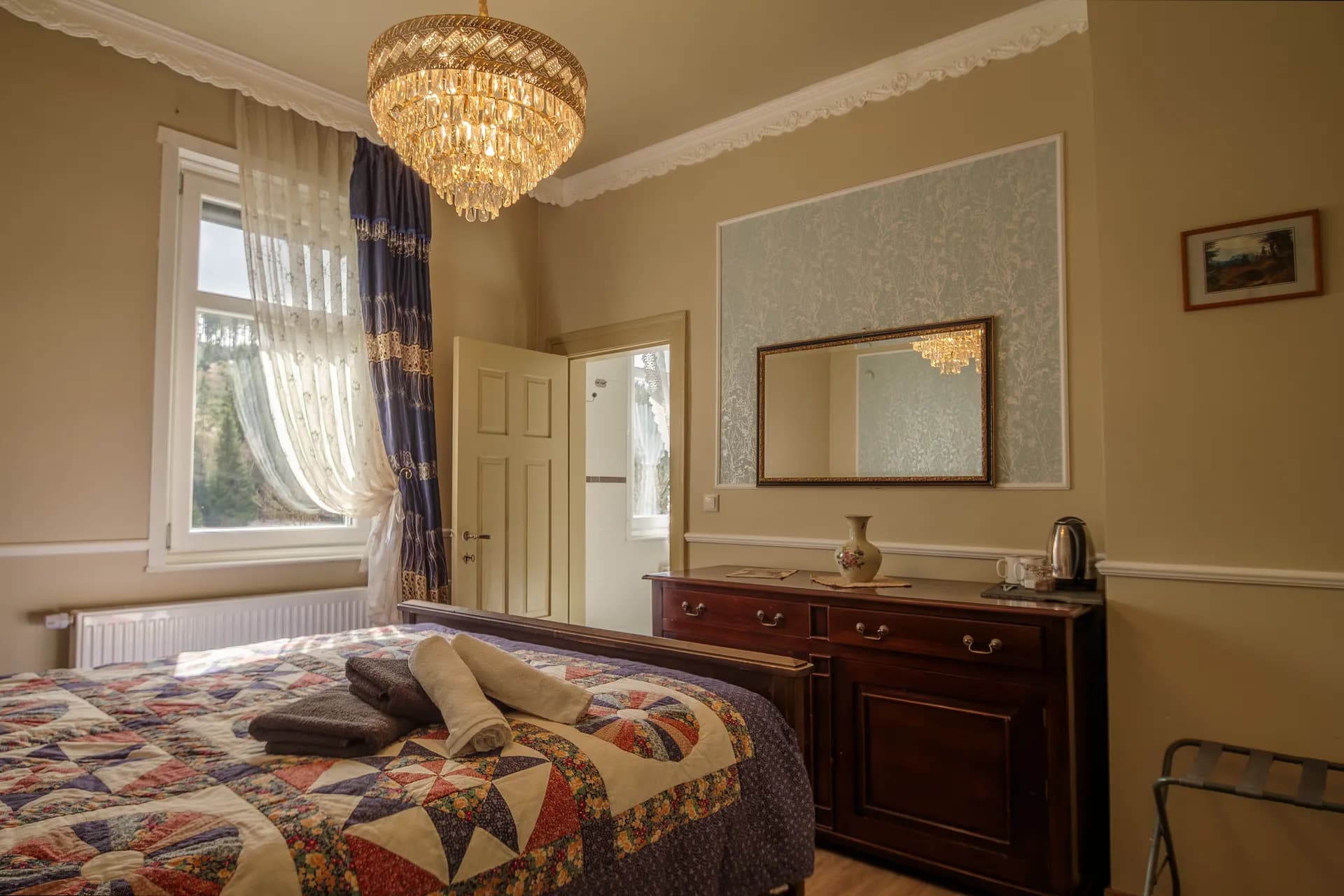 Gemütliches Hotelzimmer mit großem Doppelbett samt Patchwork-Tagesdecke, edler Holzkommode mit Wandspiegel, Kaffeestation, Kristalllüster und Zugang zum Bad.