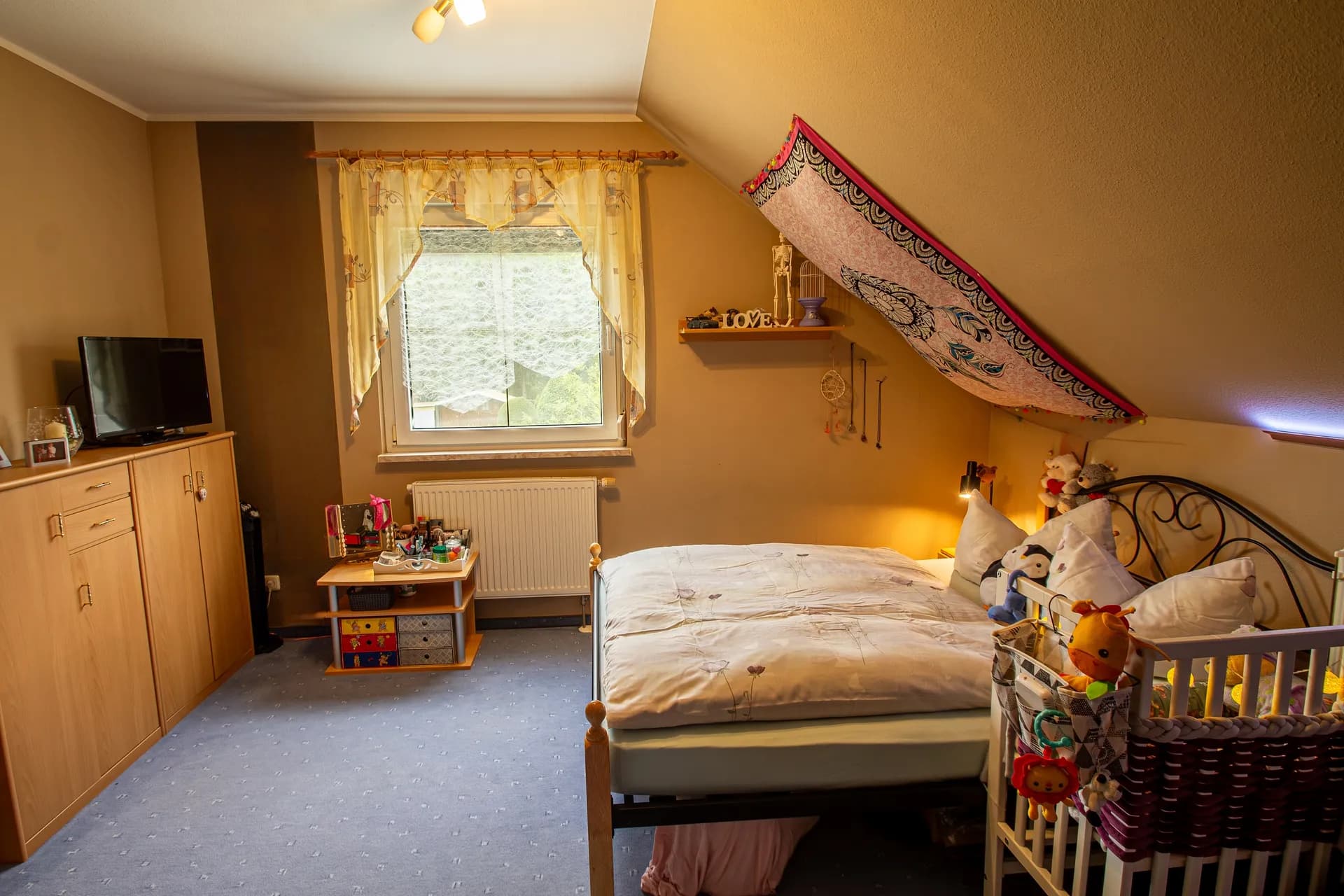 Helles Schlafzimmer im Dachgeschoss mit großem Fenster, Schrankelementen und Platz für Kinderbett.