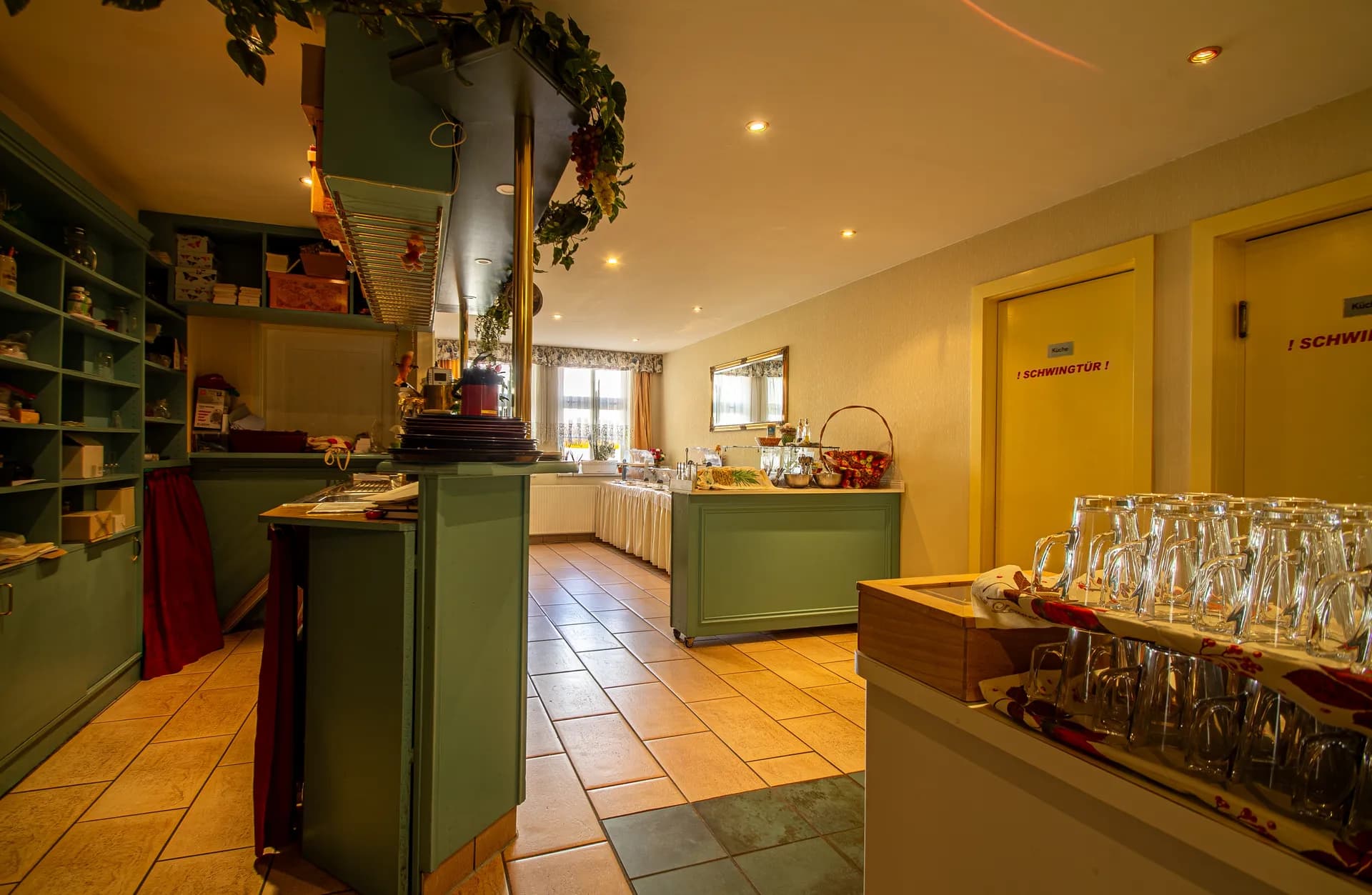 Gastronomischer Service- und Ausschankbereich mit Gläserstation, Regaleinbauten, dekorativer Hängedekoration, Buffet-Anrichte im Hintergrund und Zugang über Pendeltüren („Schwingtür“).