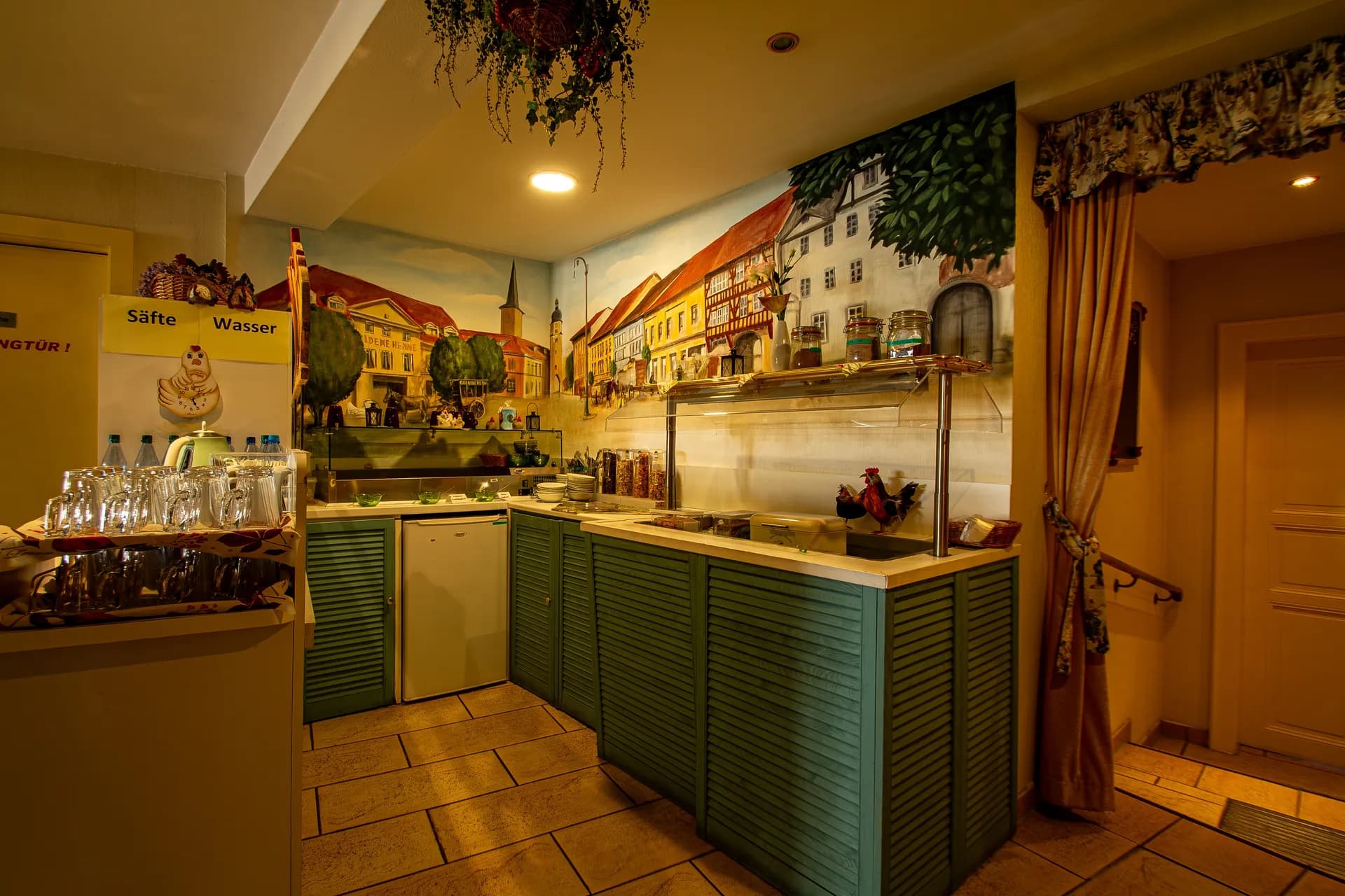Frühstücksbuffet-Bereich des Hotels mit mintgrüner Anrichte, integrierter Kühltheke, Getränkestation („Säfte / Wasser“), detailreicher Wandmalerei mit historischer Stadtansicht und Fliesenboden.