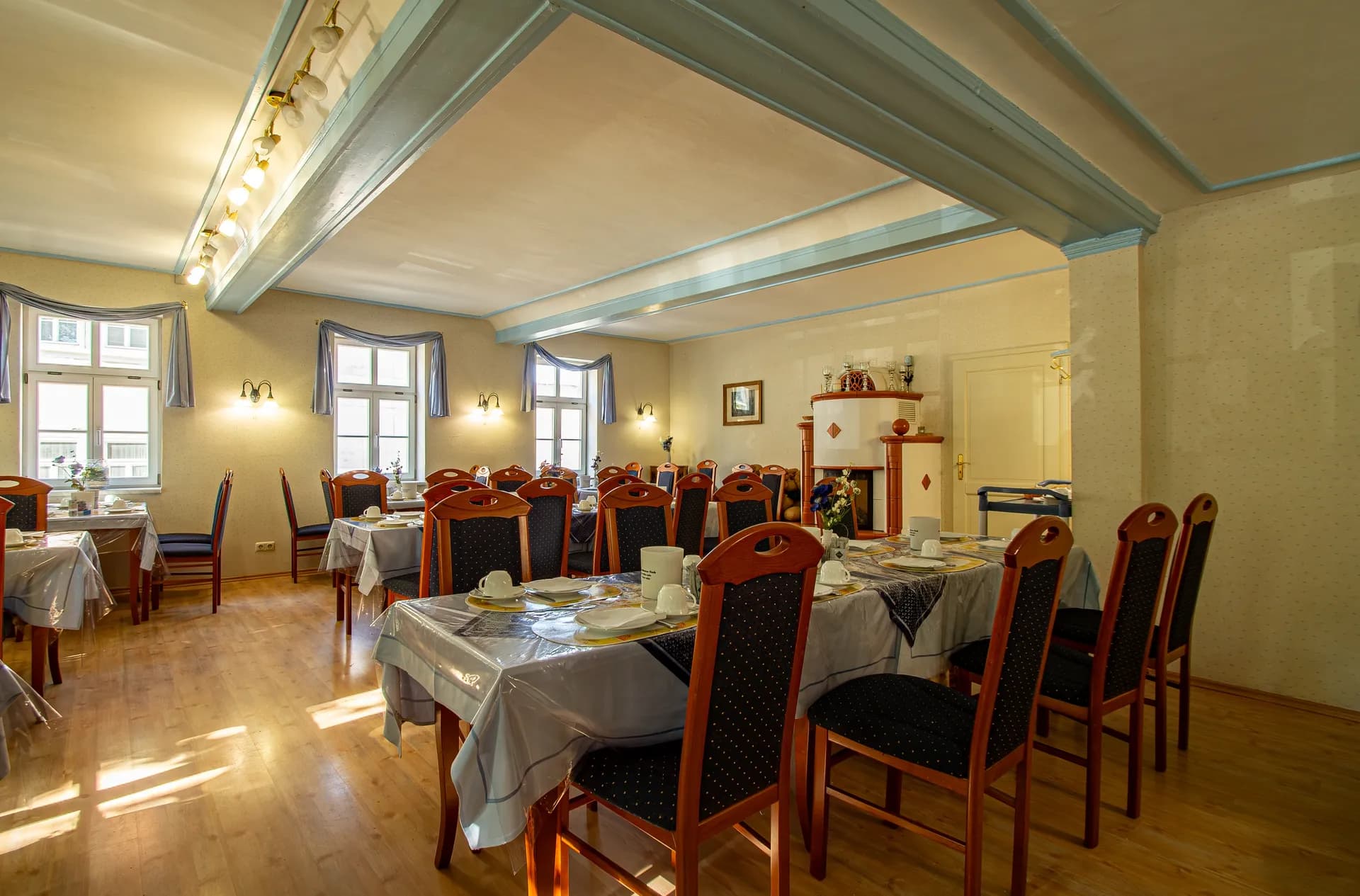Einladender Restaurant- und Veranstaltungsraum mit festlich arrangierter Festtafel, Deckenbeleuchtungsportal, Holzboden und traditionellem weißen Kachelofen im Hintergrund