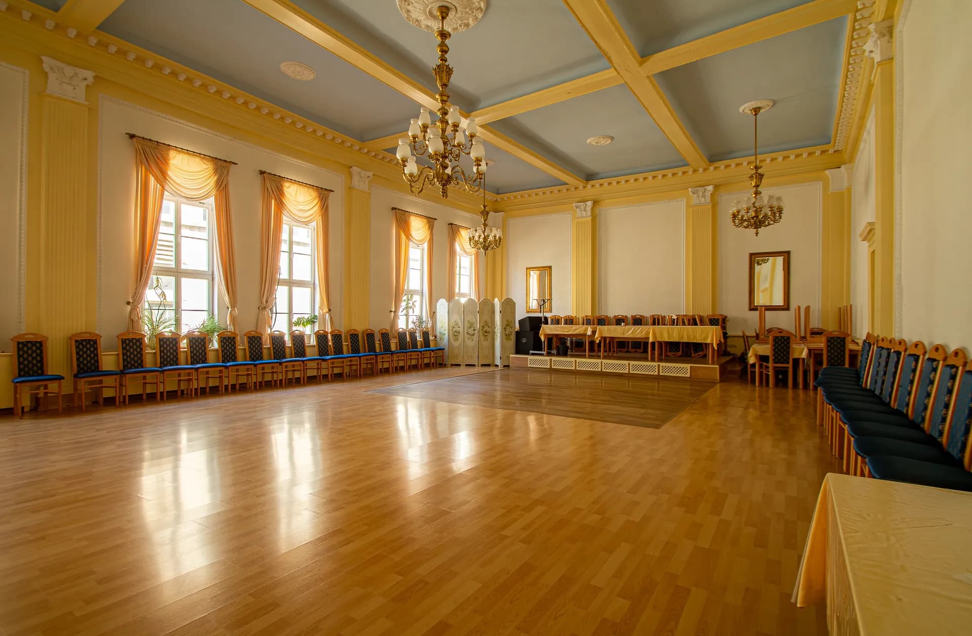 Repräsentativer historischer Ballsaal mit hellblau abgesetzter Kassettendecke, stuckierten Pilastern, prunkvollen Kronleuchtern, Parkett-Tanzfläche und gereihter Bestuhlung an den Wänden.