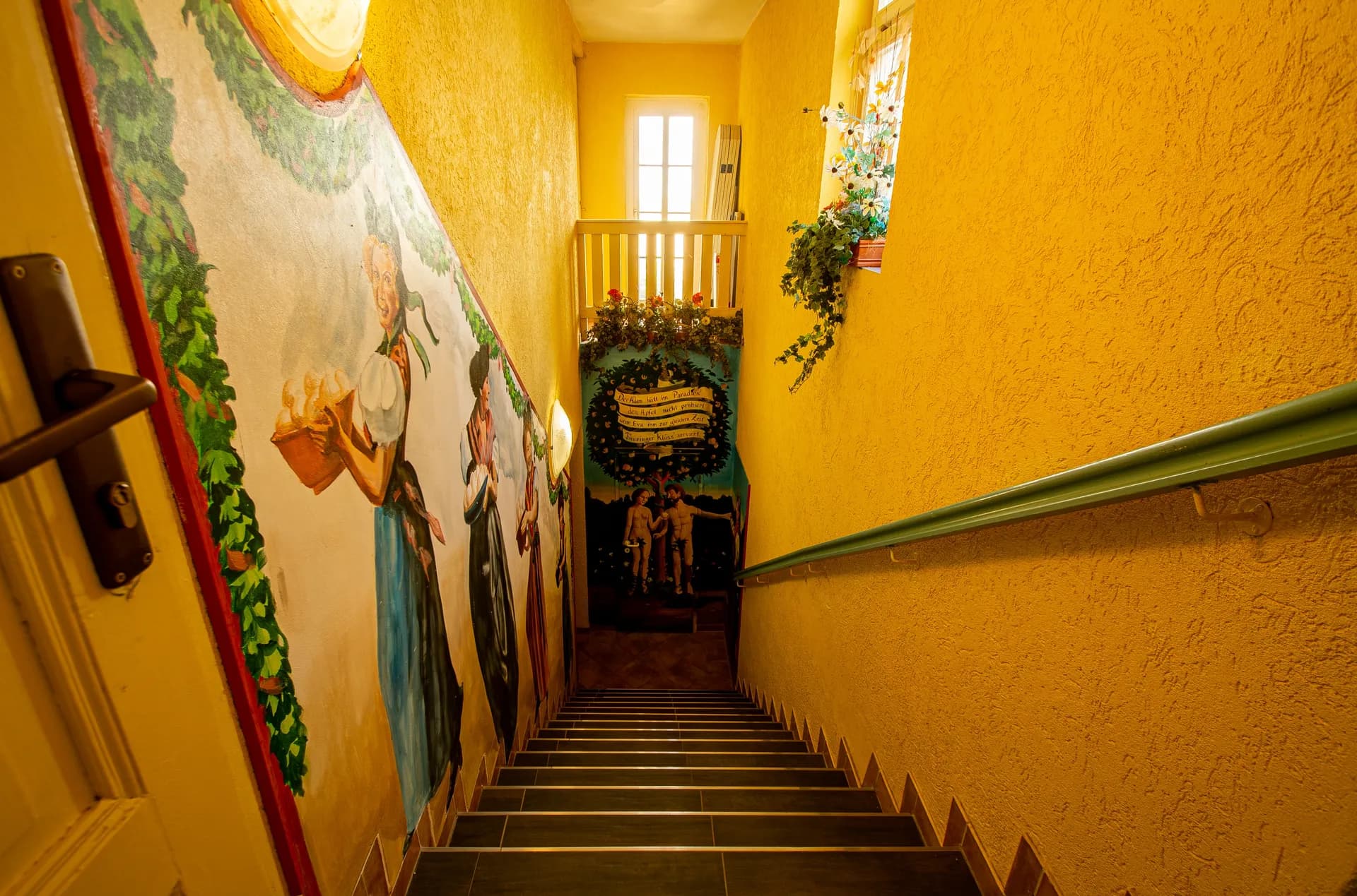 Treppenabgang mit ockerfarbenem Strukturputz, handgemalter Wandmalerei mit traditionellen Trachtenmotiven, grünem Handlauf und tageslichtdurchflutetem oberen Treppenpodest.