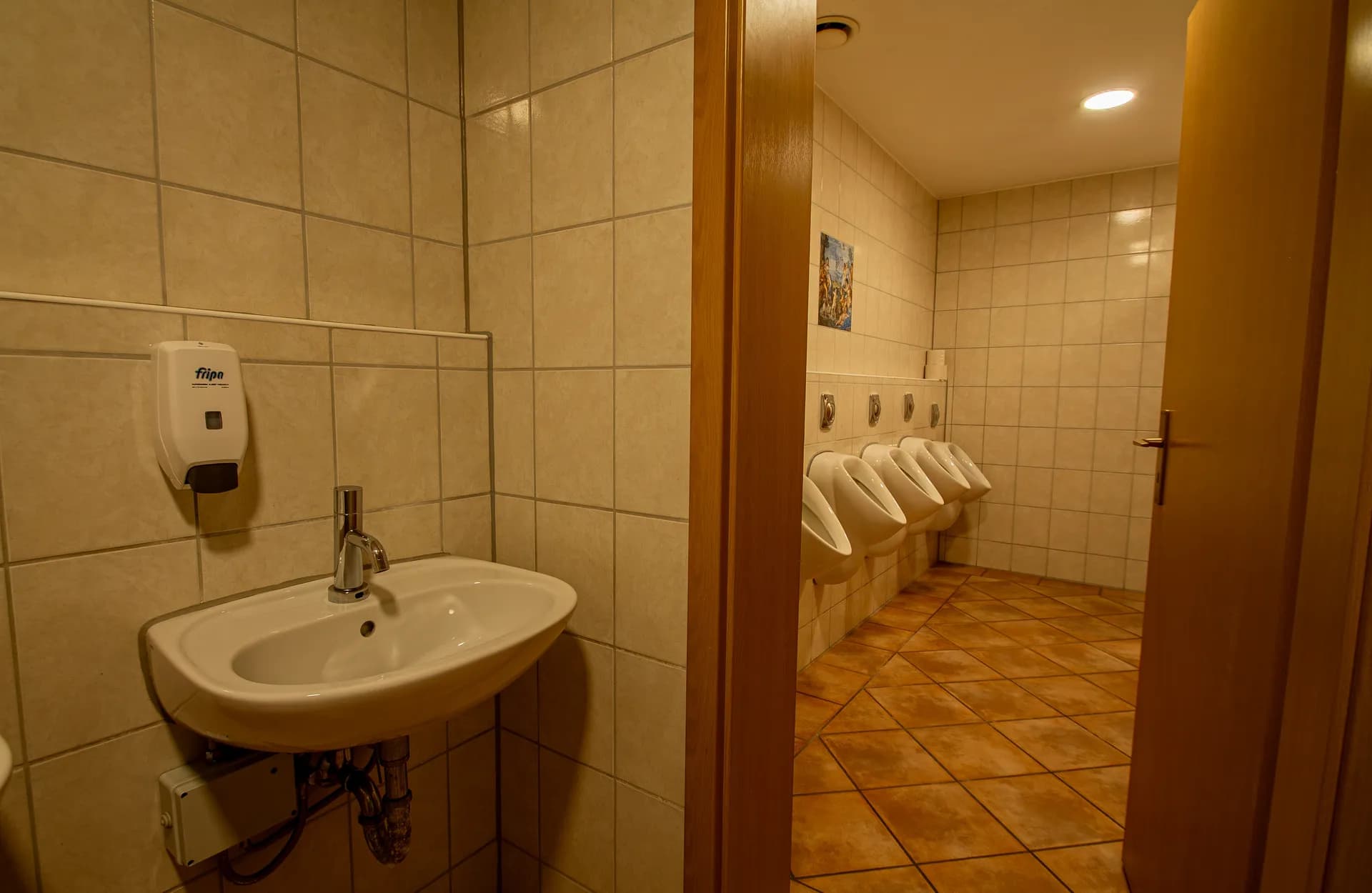 Geräumiger Sanitär-Vorraum mit drei Handwaschbecken samt Desinfektionsmittelspendern, wandmontiertem Wickeltisch, Vintage-Spiegel und Terrakotta-Bodenfliesen.