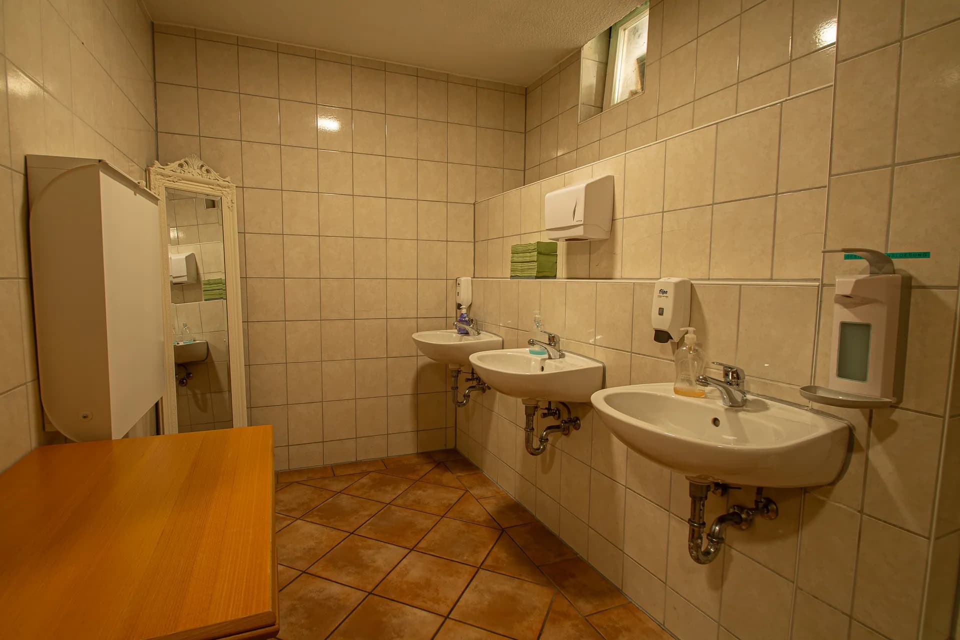 Gäste-Sanitärbereich (Herren-WC) im Gastronomiebereich mit Wandwaschbecken, Seifenspender, Fliesenbelag und Durchblick zur Urinal-Anlage.