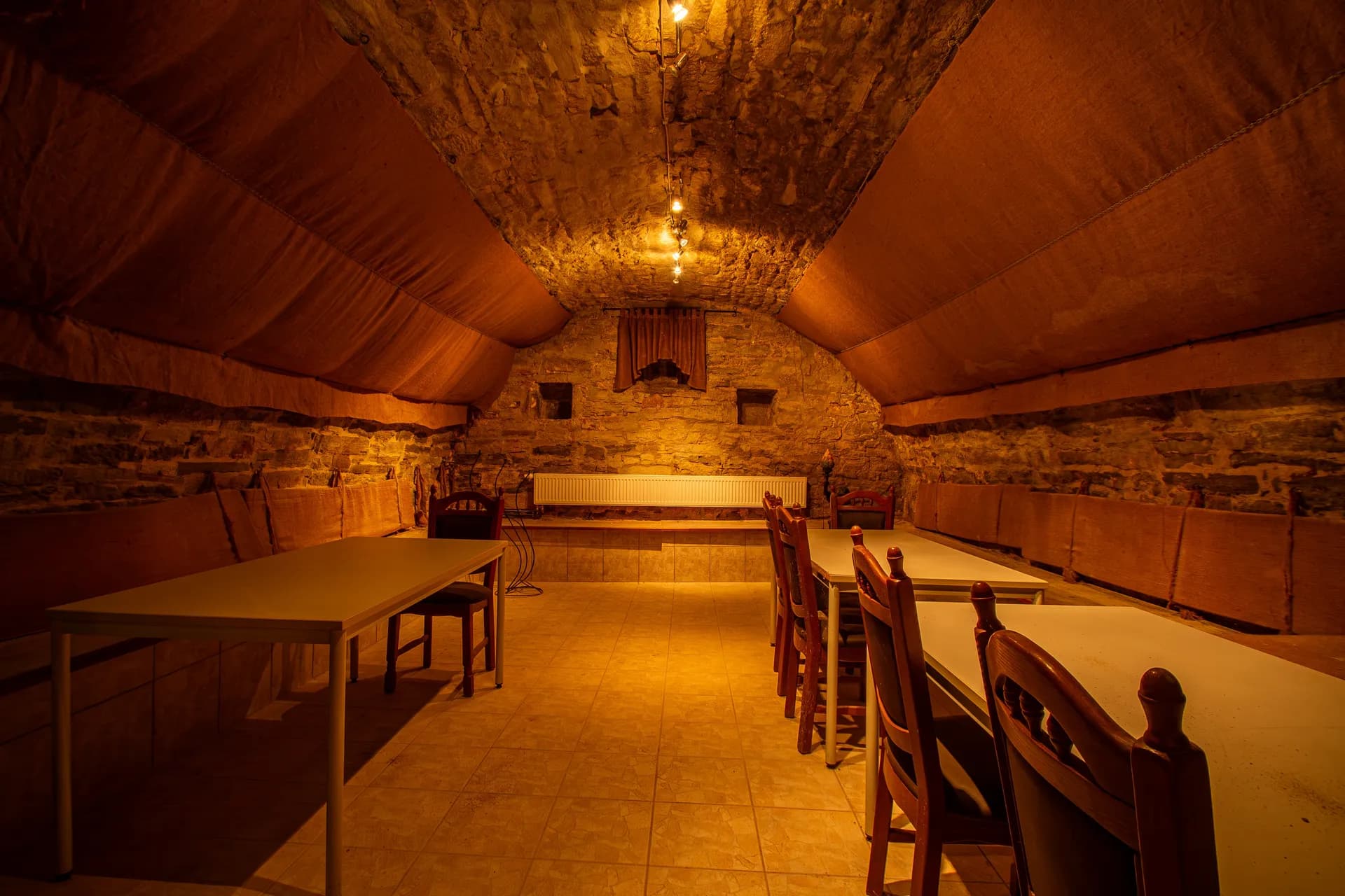 Rustikaler historischer Gewölbekeller / Weinkeller mit Natursteingewölbe, stoffbespannten Deckensegeln, warmer Beleuchtung, Holztischen und umlaufenden Sitzbänken.