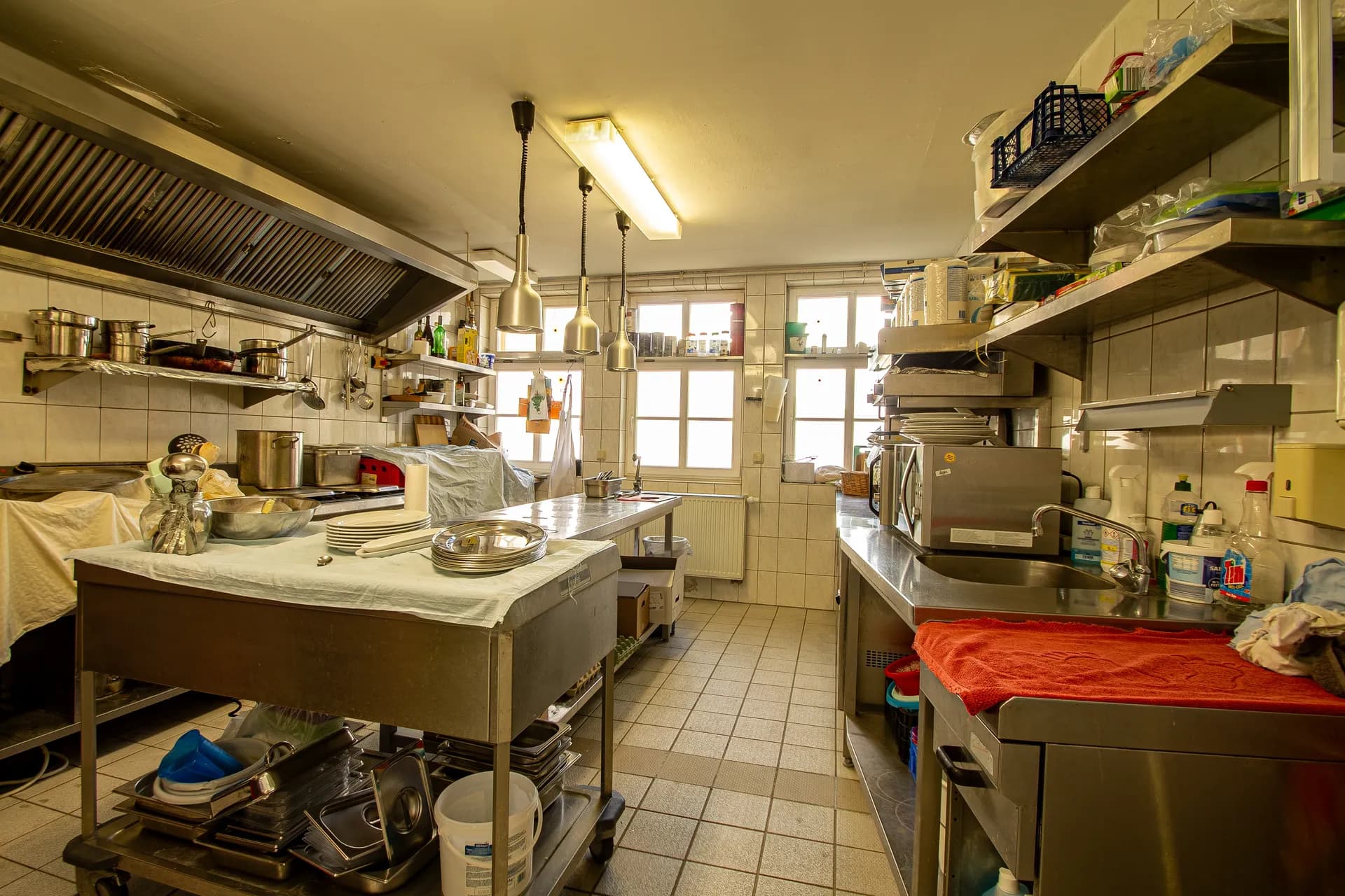 Gewerbliche Großküche des Restaurantbetriebs mit professionellen Edelstahl-Arbeitsflächen, Dunstabzugshaube, Spülbereich, Regalen und gefliesten Wänden.