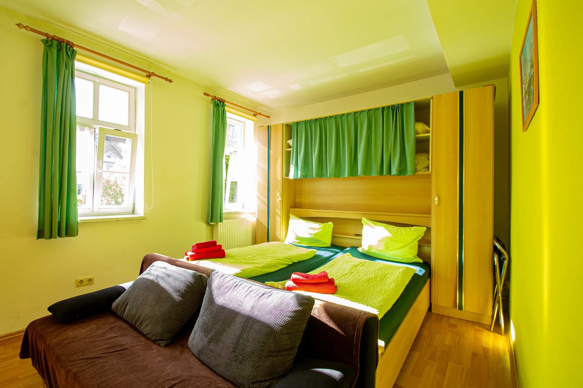 Sonniges Gästezimmer mit hellen Holz-Einbaumöbeln, Doppelbett mit lindgrüner Bettwäsche, vorgelagertem Schlafsofa und zwei Fenstern mit passenden Vorhängen.