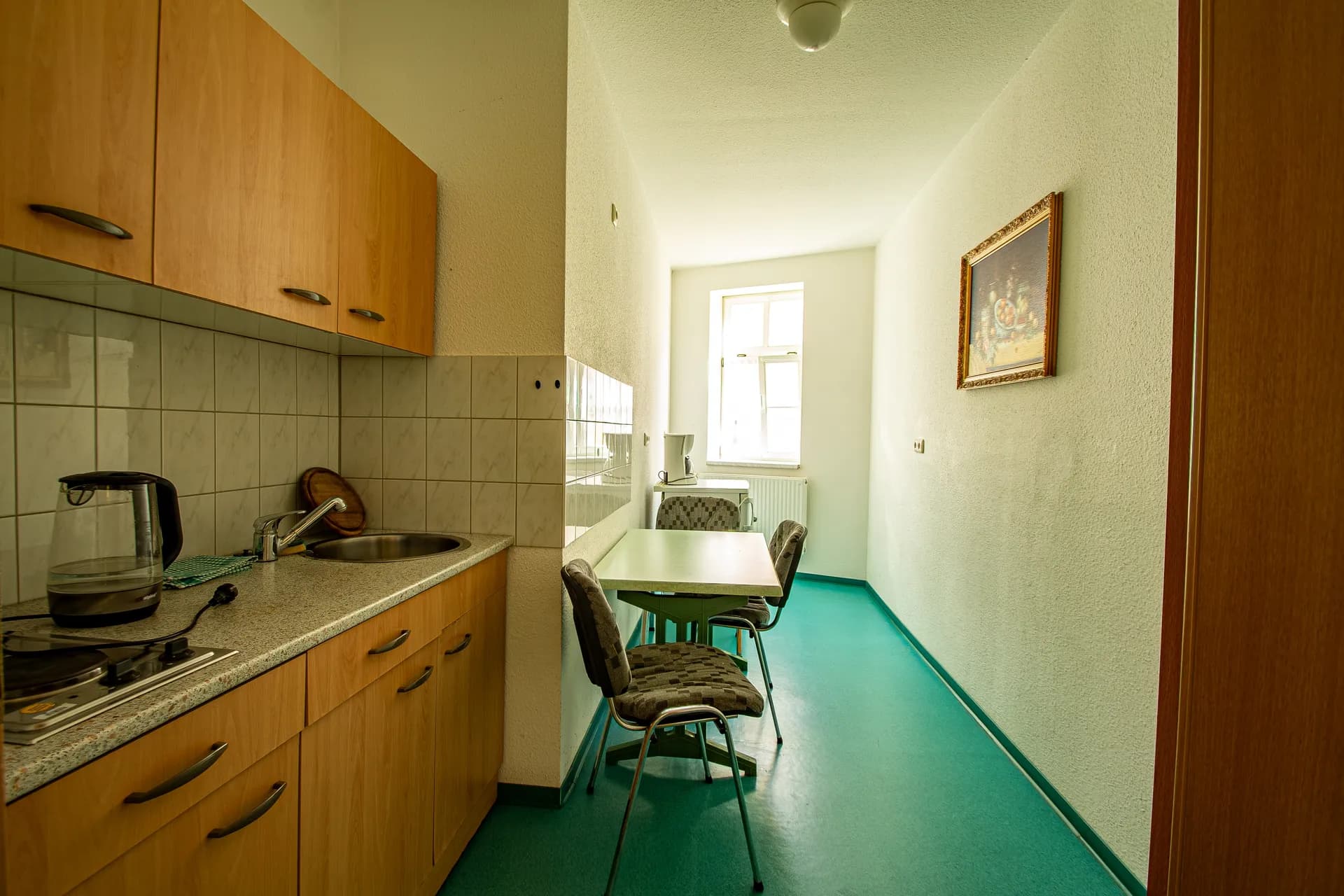 Länglich geschnittene Apartmentküche mit Einbauzeile aus Holz, Spülbecken, Kochfeld, kleinem Esstisch mit Stühlen, türkisfarbenem Bodenbelag und Fenster an der Stirnseite.