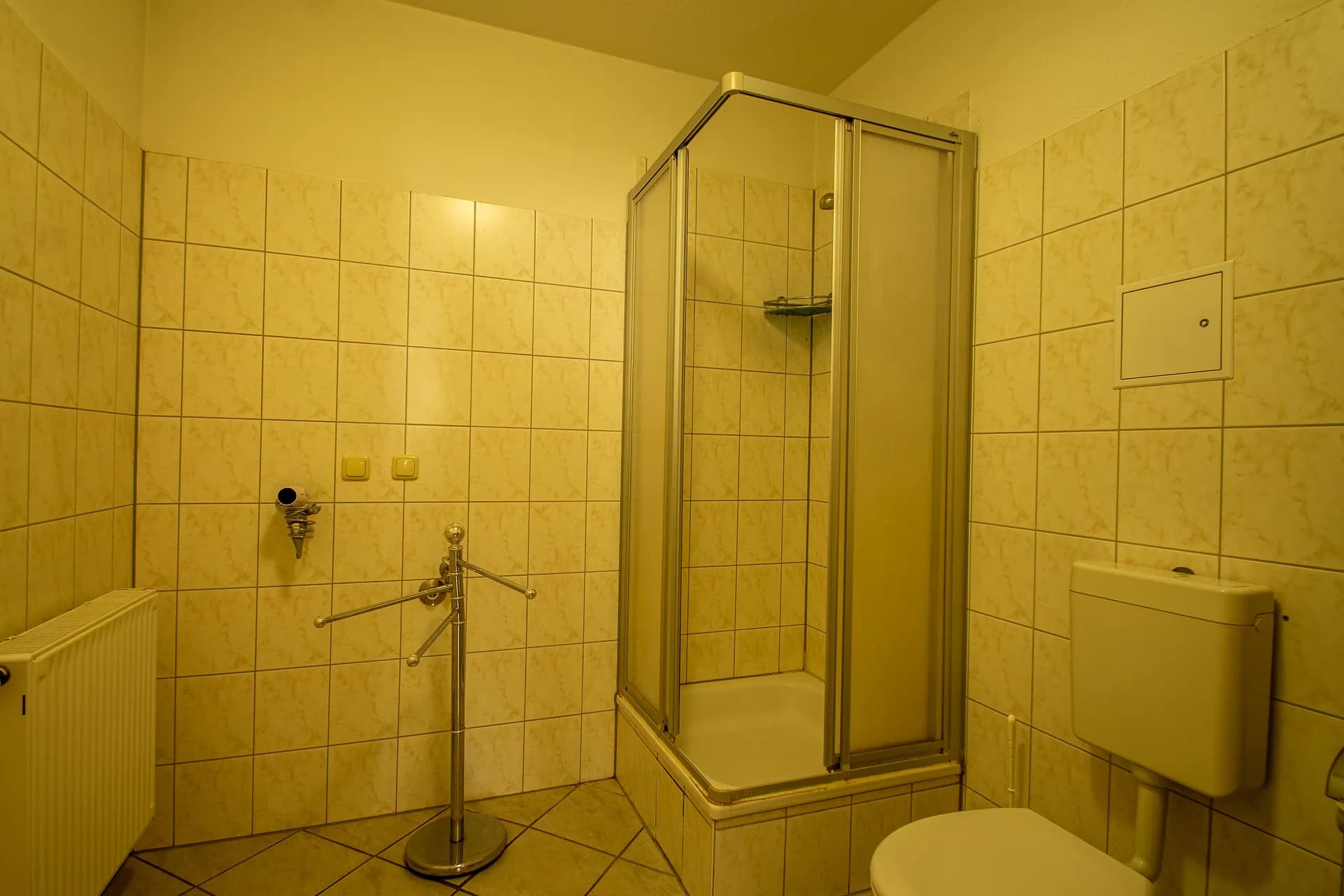 Kompaktes Badezimmer mit beiger Verfliesung, Eckduschkabine samt Schiebetüren, Stand-WC, freistehendem Handtuchhalter und Heizkörper.