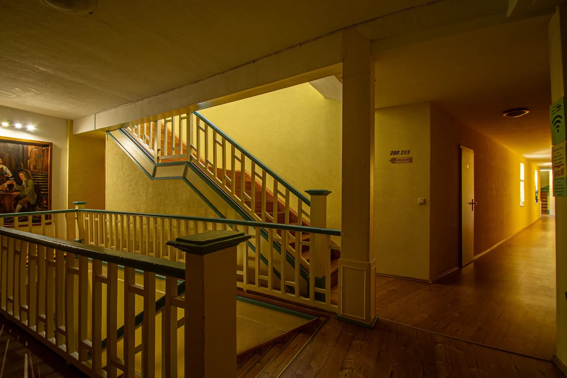 Großzügiger Etagenflur und Treppenabsatz des Hotels mit Holzgeländer, beleuchtetem Wandgemälde, Zimmerwegweiser (200–211) und stimmungsvoller Beleuchtung.