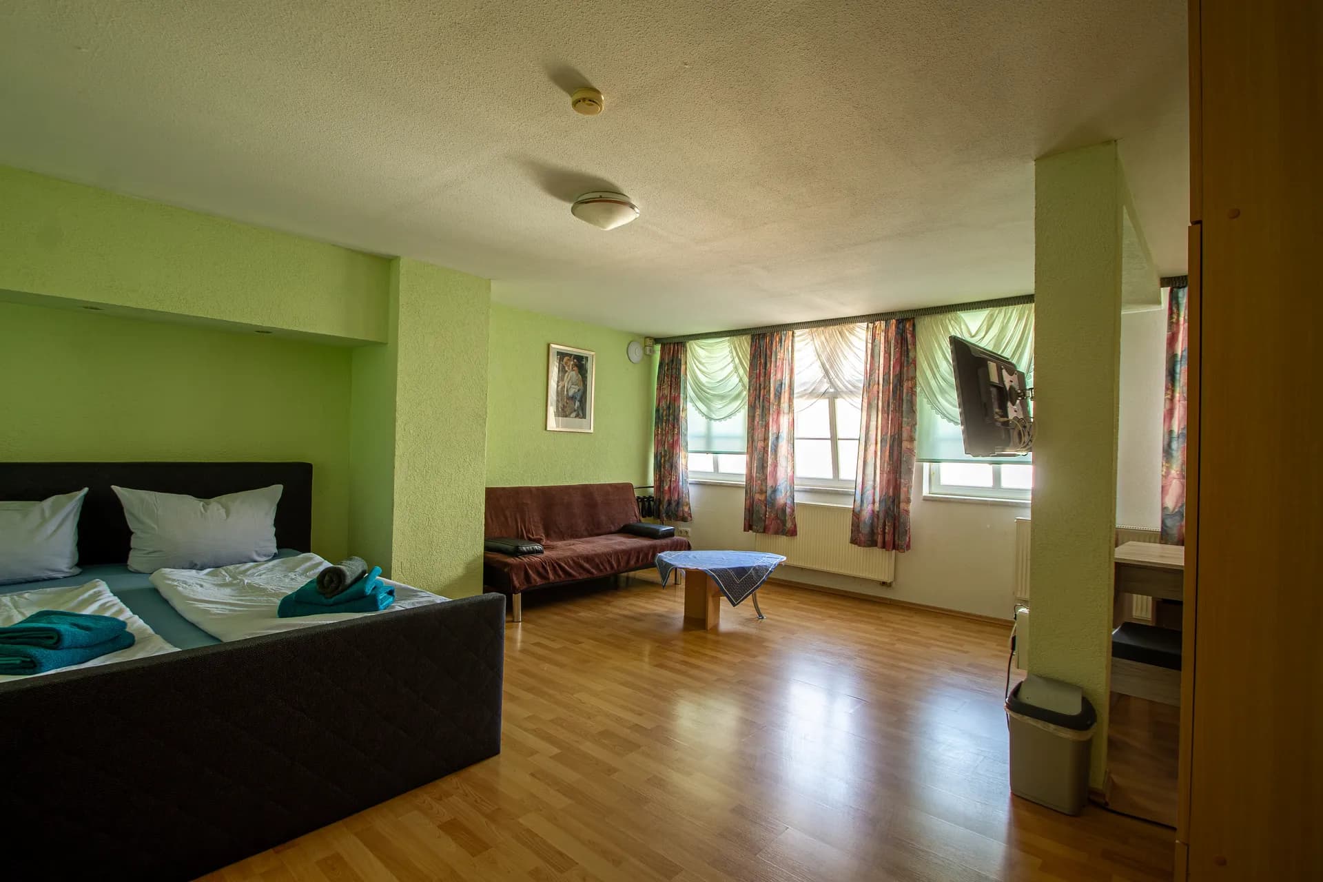 Geräumiges Studio- oder Hotelzimmer in Hellgrün mit gepolstertem Doppelbett, Sitzecke samt Sofa und Couchtisch, Laminatboden, wandmontiertem TV und breiter Fensterfront.