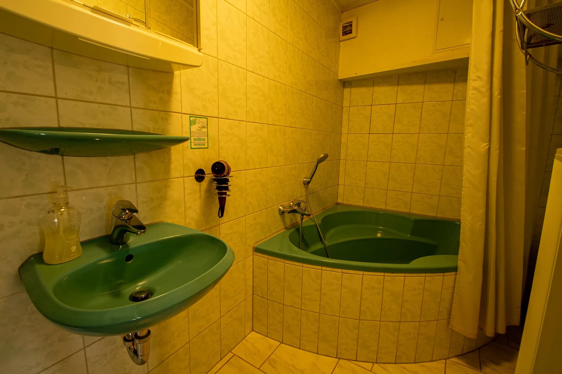 Badezimmer mit markanter smaragdgrüner Sanitärausstattung, integrierter Eckbadewanne samt Duschvorhang, passendem Waschbecken, Wandregal und Haartrockner.