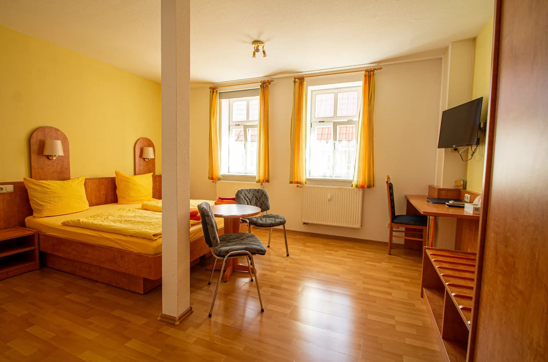 Helles Hotelzimmer in sonnigen Gelbtönen mit Holz-Doppelbett, kleiner Sitzecke samt Rundtisch, Schreibtisch mit TV, Kofferablage und Laminatboden.