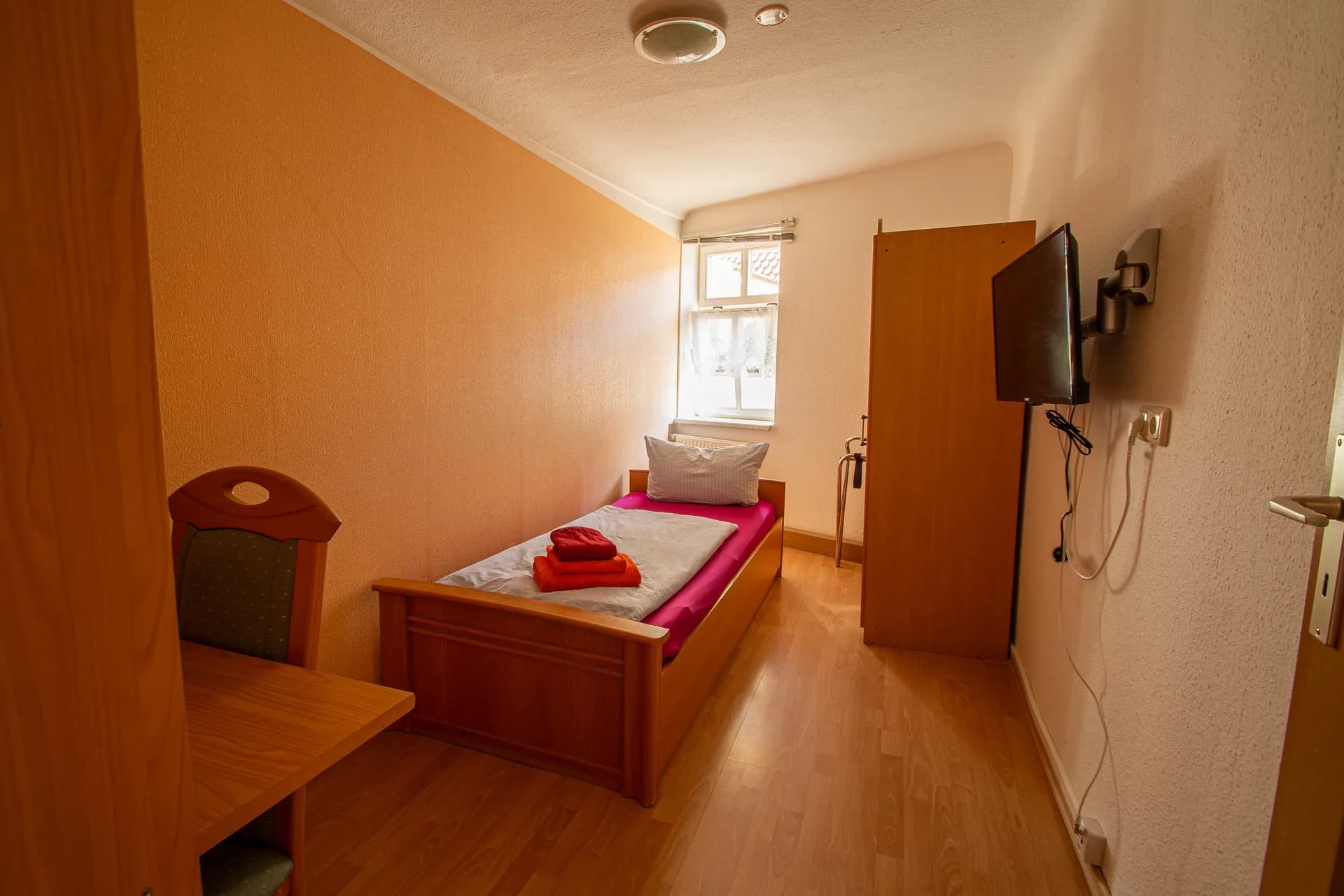 Gemütliches Einzelzimmer mit Massivholz-Einzelbett, Kleiderschrank, kompaktem Schreibtisch samt Stuhl, wandmontiertem Flachbildfernseher und Tageslichtfenster.