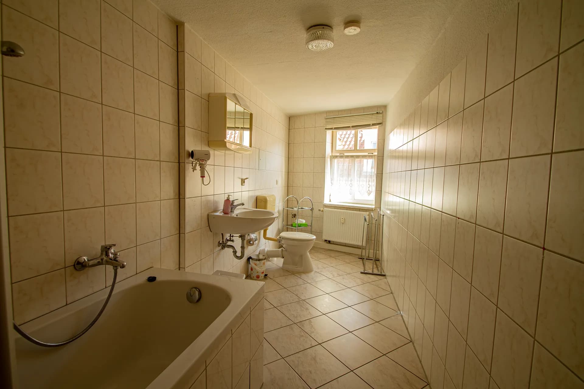Großzügiges Badezimmer mit heller Wandverfliesung, Einbau-Badewanne, Waschtisch mit Spiegelschrank, Stand-WC und stirnseitigem Fenster.