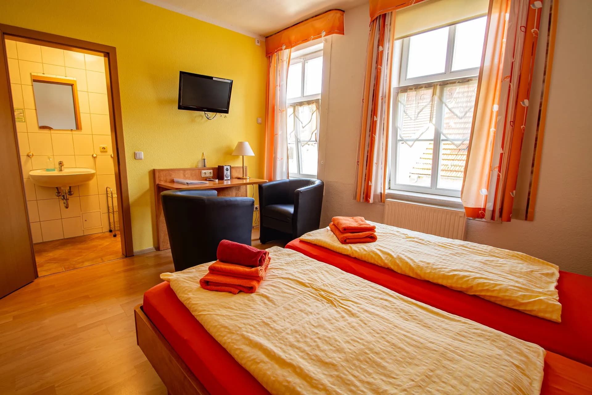 Farbenfrohes Doppelzimmer mit sonnengelber Wandgestaltung, zwei Betten in orange-gelber Bettwäsche, ledernen Clubsitzern, TV-Wand und direktem Zugang zum eigenen Duschbad.  IMG_7717-HDR.jpg