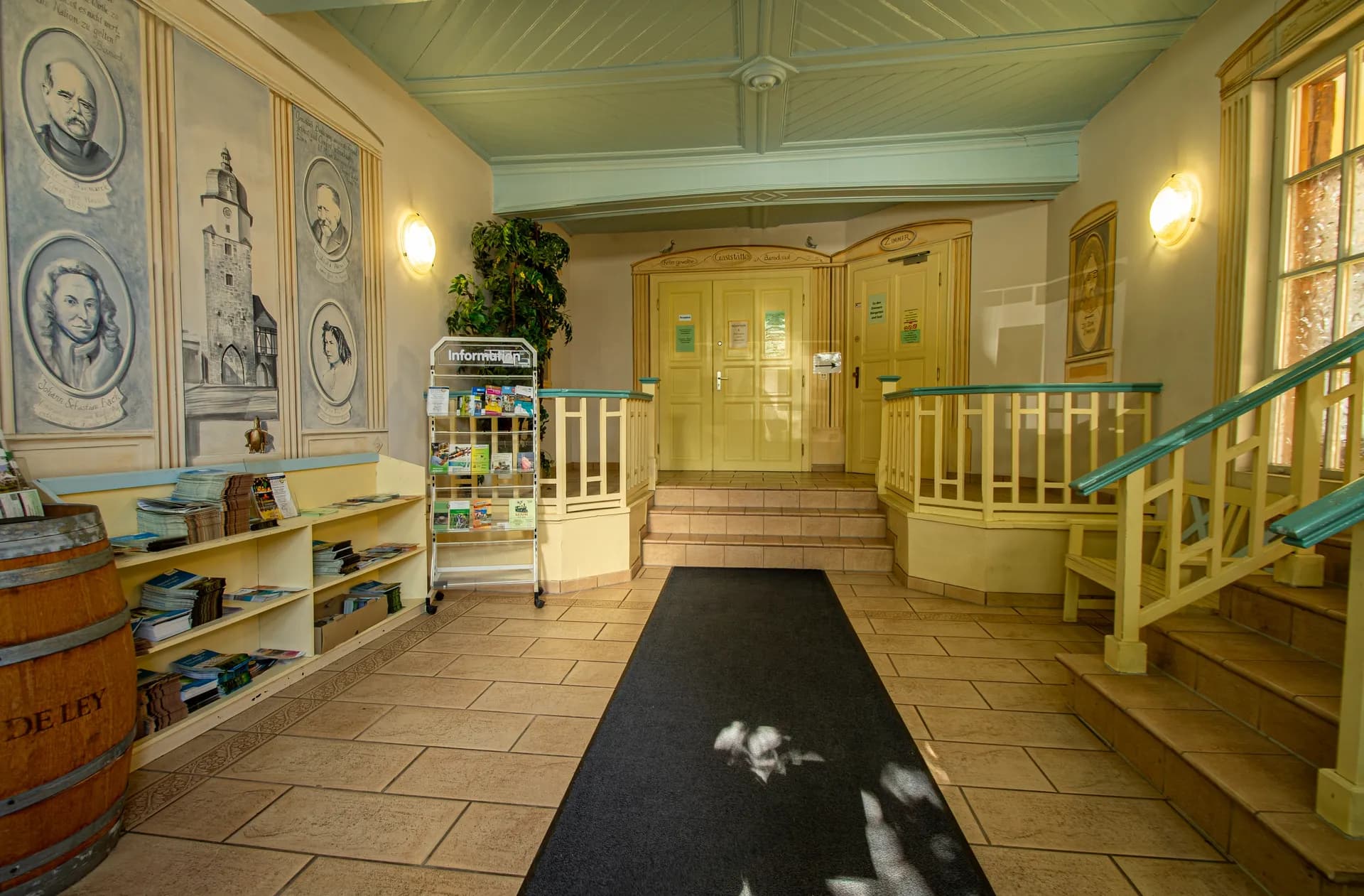 Weitläufiges Hotelfoyer mit mintgrüner Holzkassettendecke, historischer Wandmalerei mit Porträts, Broschürenständern, dekorativem Weinfass und zentralem Teppichläufer.