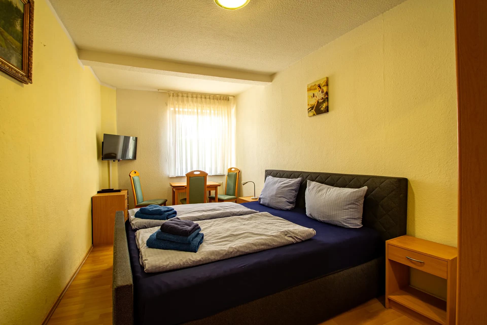 Helles Doppelzimmer in Gelbtönen mit dunkelblau gepolstertem Doppelbett, Nachttisch, Sitzecke mit Tisch und Stühlen, wandmontiertem TV und Laminatboden.