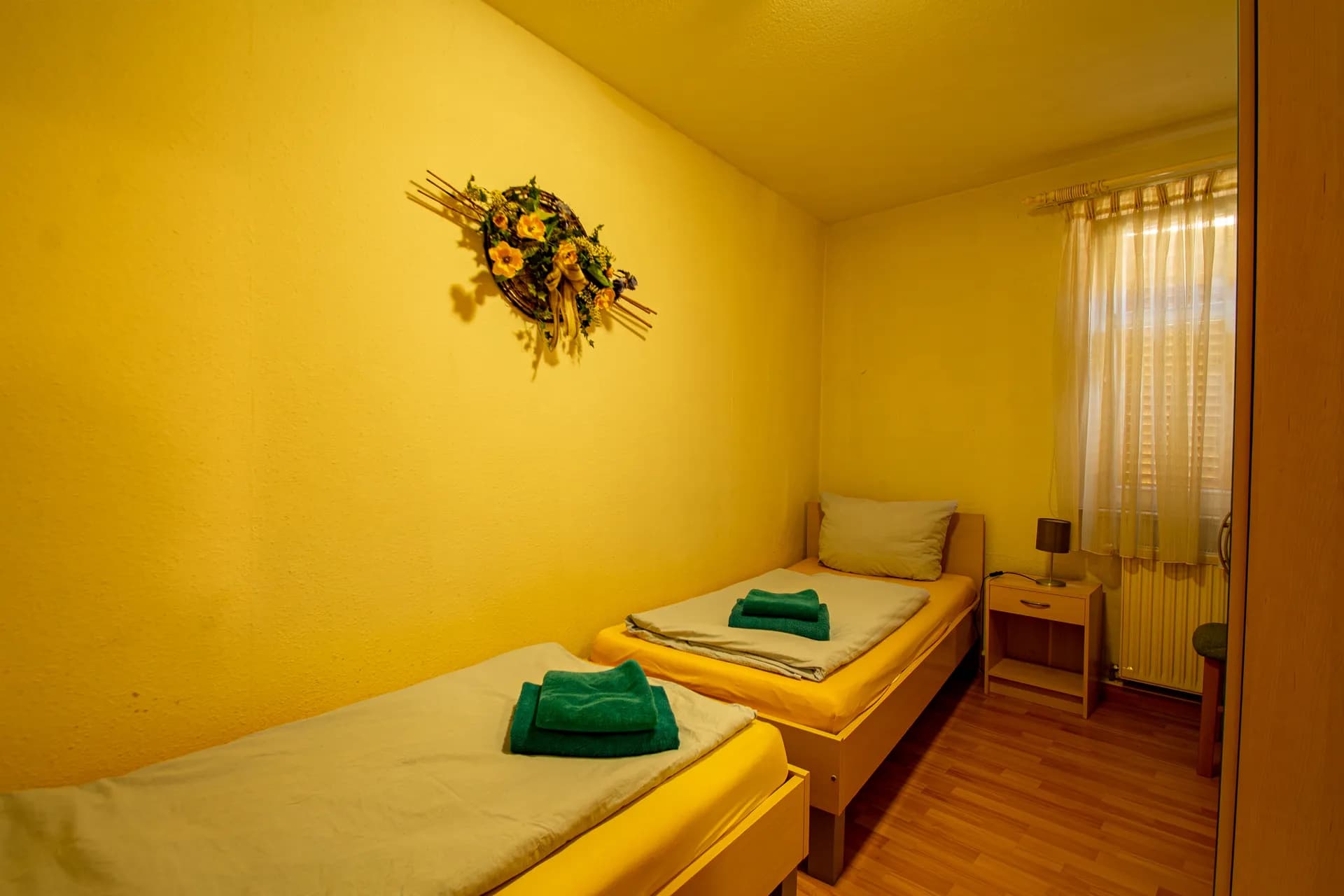 Gemütliches Zweibettzimmer mit zwei getrennt stehenden Holz-Einzelbetten, Nachttisch mit Leselampe, floralem Wandschmuck und Fenster mit zarter Gardine.