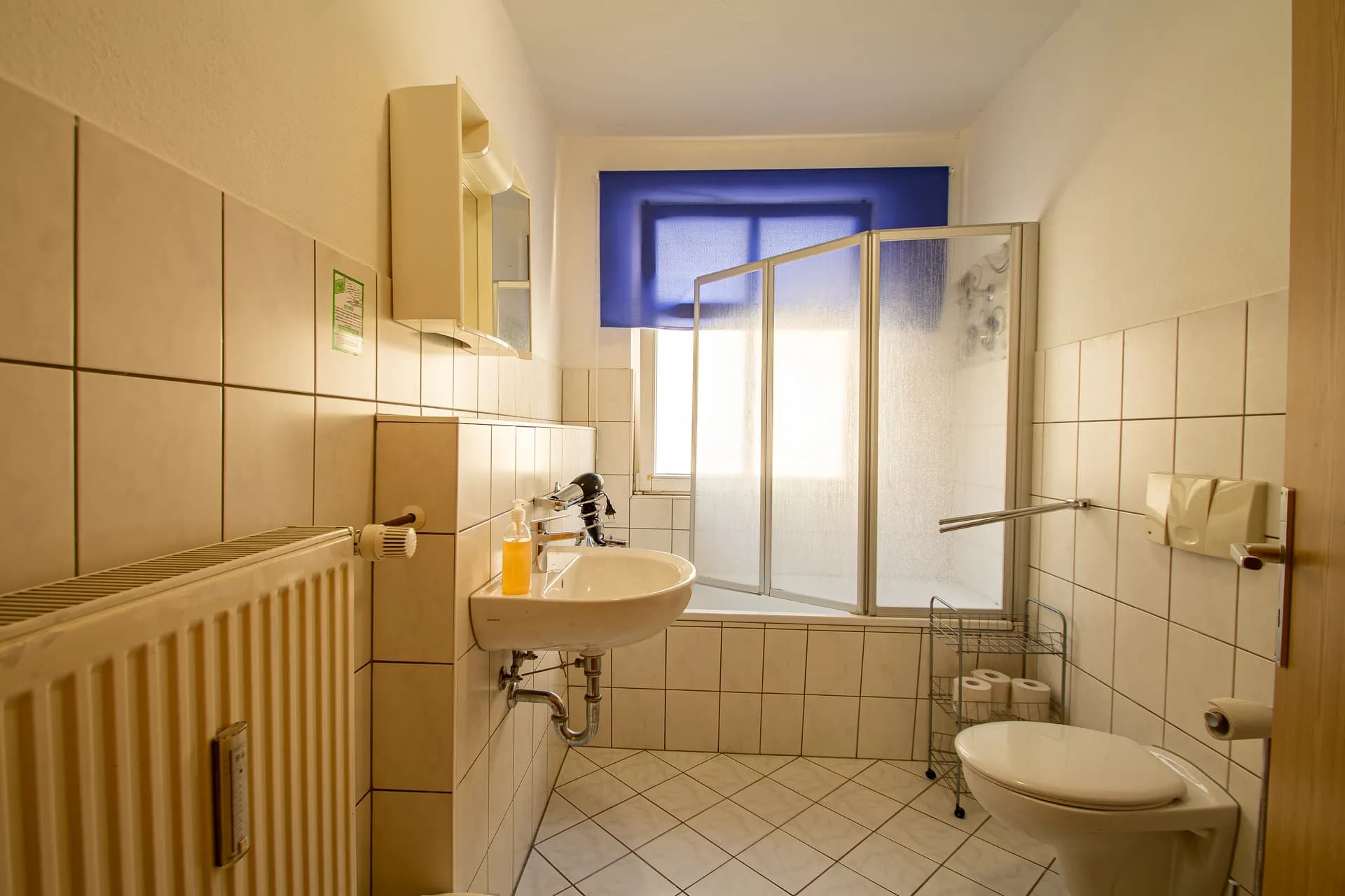 Helles Badezimmer mit diagonal verlegten Fliesen, Einbau-Badewanne samt Glas-Duschabtrennung, Waschtisch mit Haartrockner, Spiegelschrank, WC und Heizkörper.