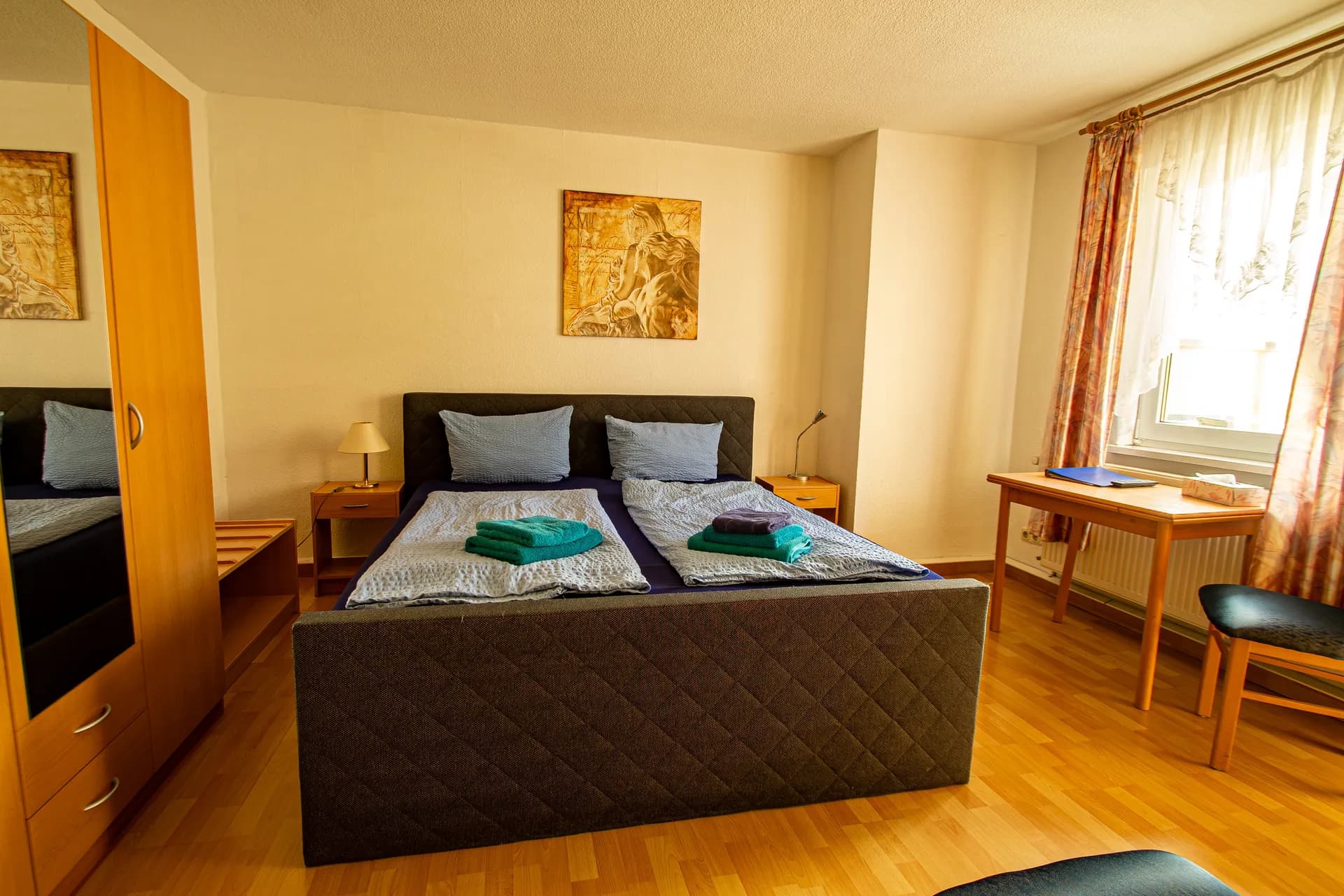 Helles Hotel-Doppelzimmer mit anthrazitfarben gepolstertem Doppelbett, Holz-Kleiderschrank samt Spiegeltür, Schreibtisch, Nachttischen und Laminatboden.