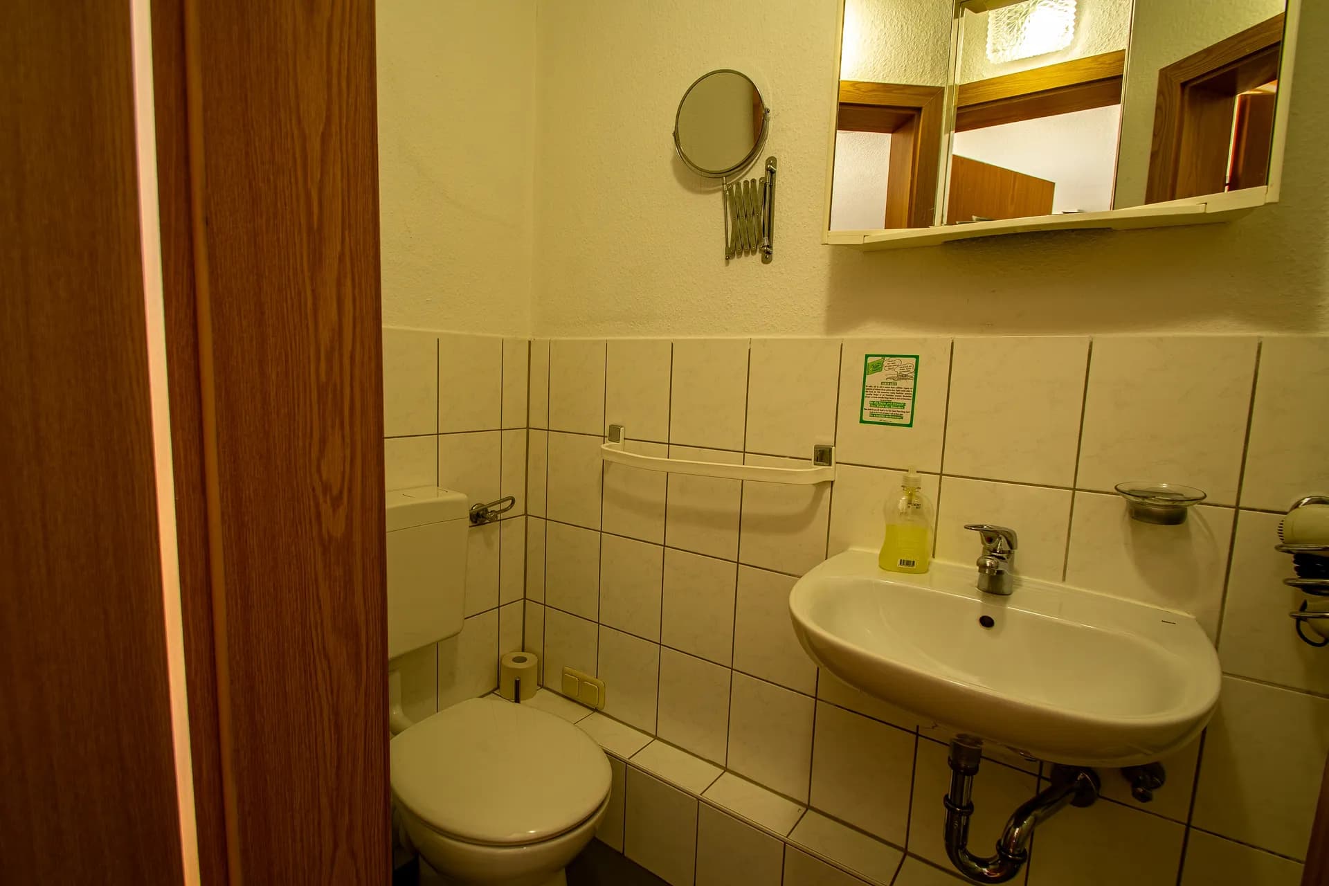 Funktionales Gäste-WC/Badezimmer mit weißer Fliesenverkleidung, Stand-WC, Wandwaschbecken samt Armatur, Kosmetikspiegel und beleuchtetem Spiegelschrank.