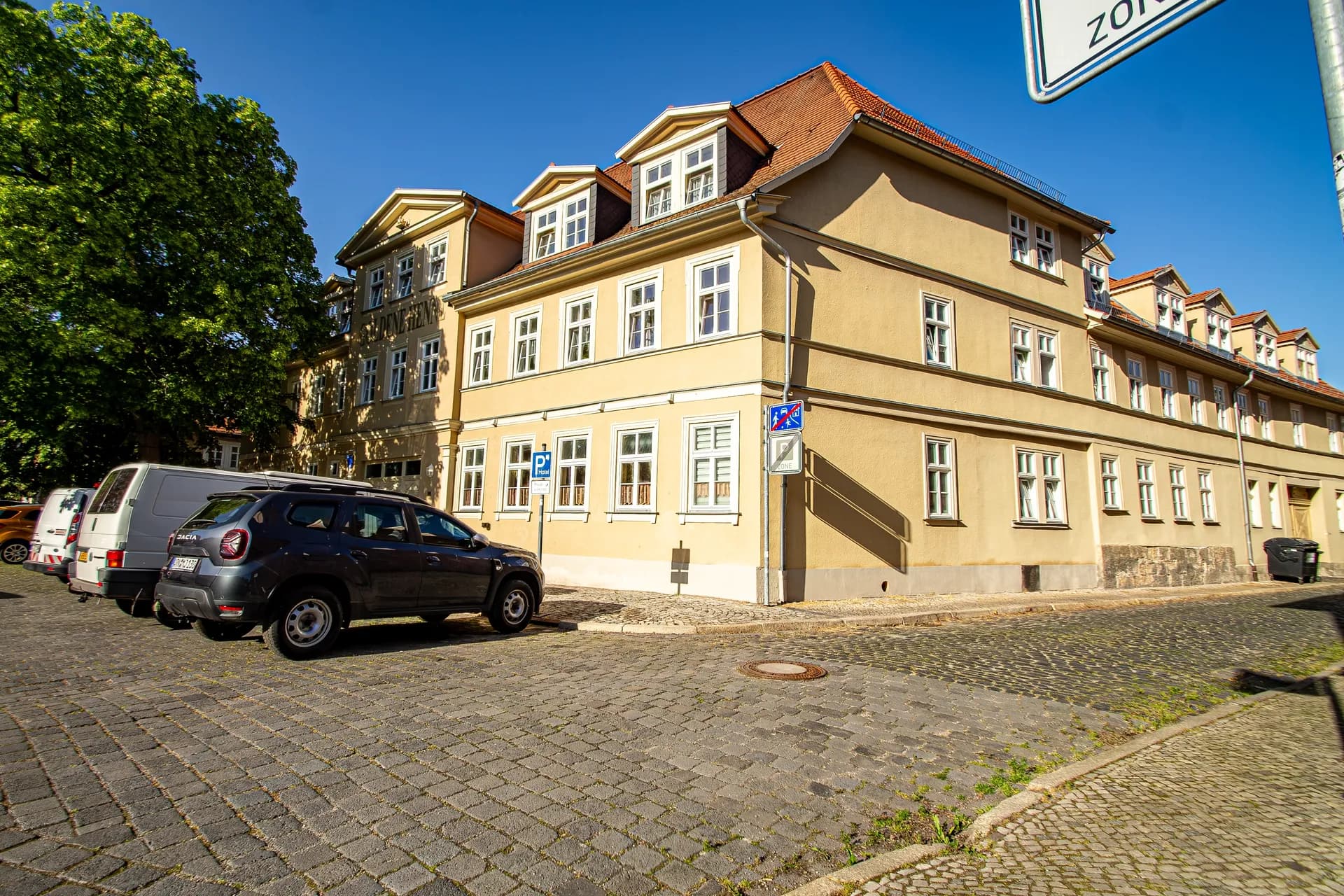 Straßen- und Platzansicht des Hotelgebäudes „Goldene Henne“ über den gepflasterten Vorplatz mit Parkmöglichkeiten, Altbaumbestand und historischer Fassadengliederung.