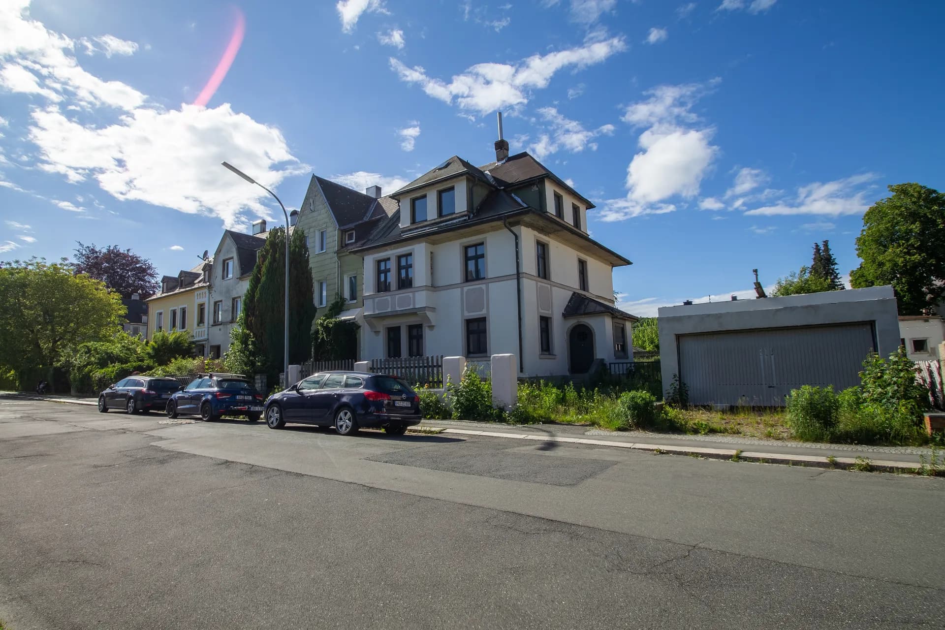 Straßenansicht und Umgebung des Mehrfamilienhauses mit Nebengebäude/Garage, Vorgarten und Parkmöglichkeiten am Straßenrand.