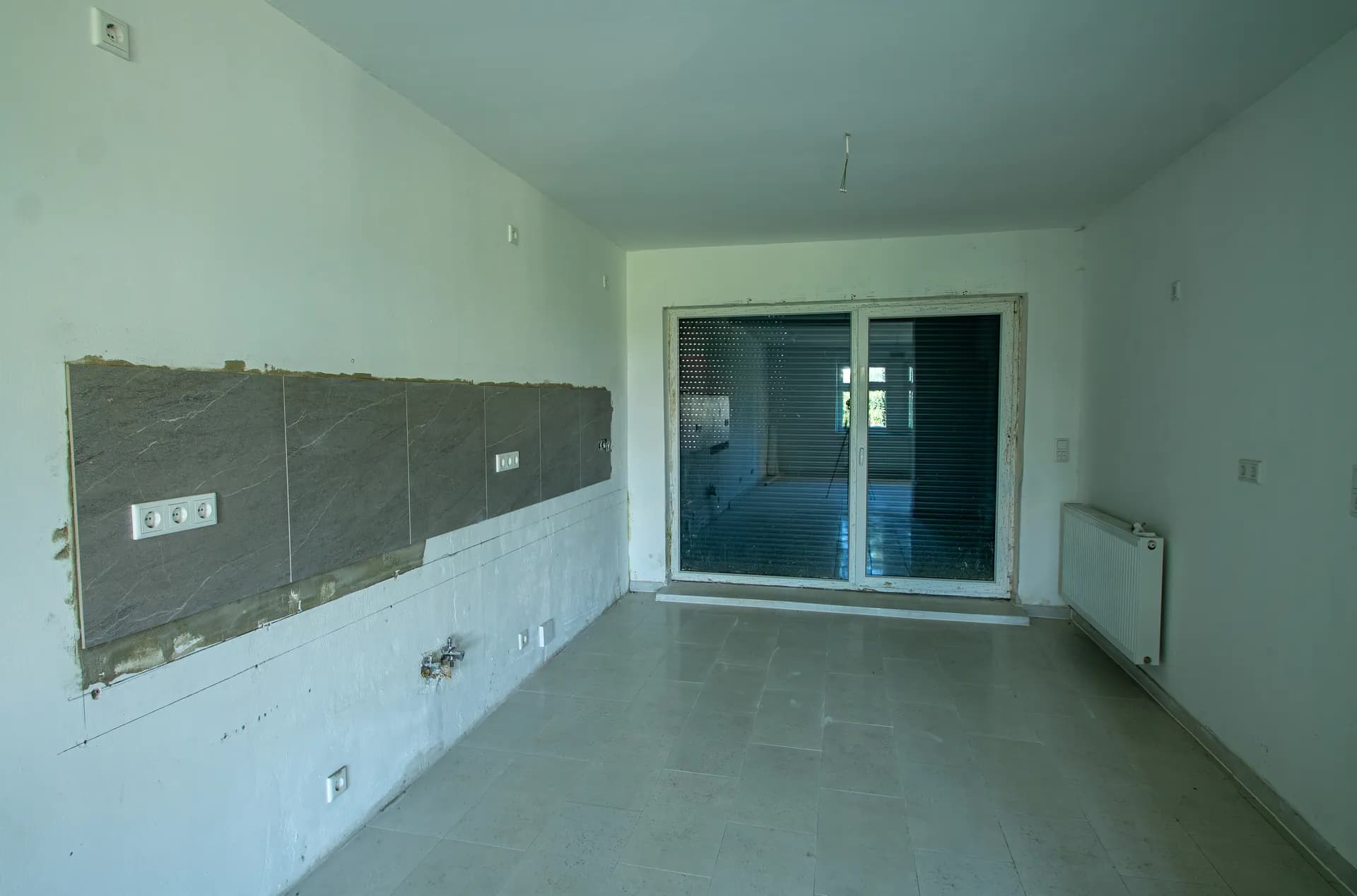 Küchen- und Essbereich mit grauem Fliesenspiegel, Elektroanschlüssen, Heizkörper und Schiebetür mit Zugang zur Terrasse.