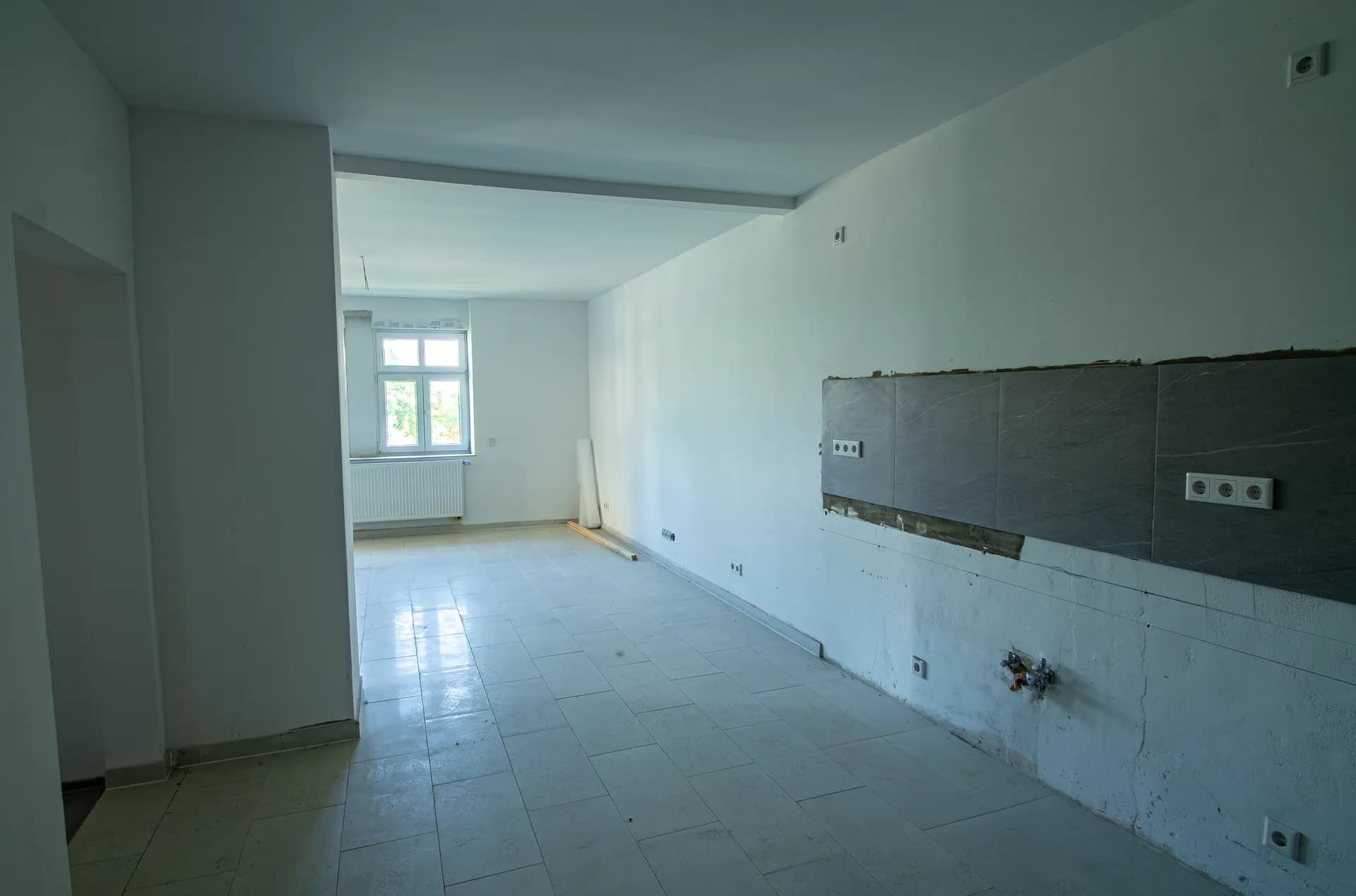 Langer Wohn- und Küchenbereich im Ausbauzustand mit Fliesenspiegel für die Einbauküche, Anschlüssen und hellem Fliesenboden.