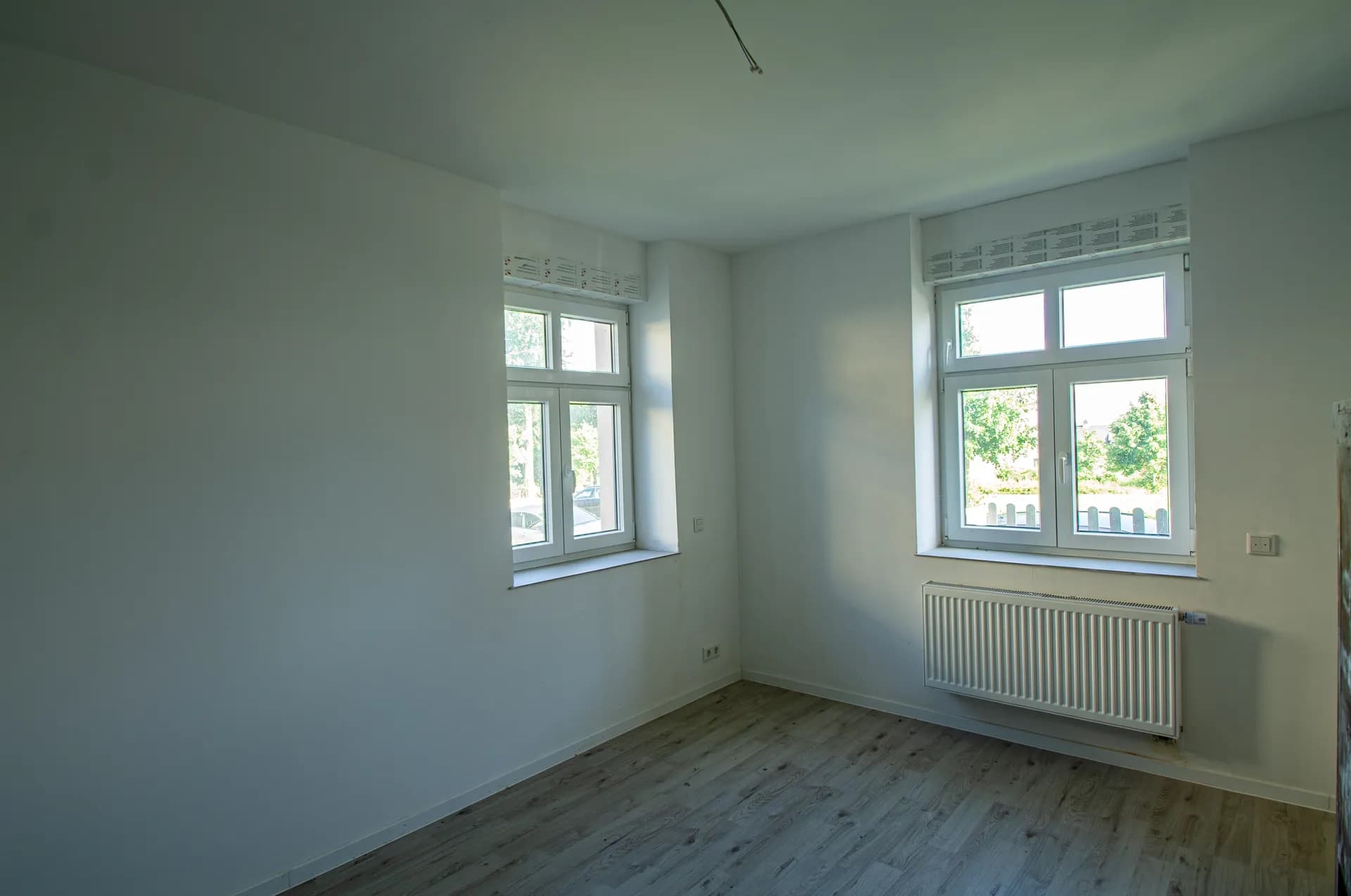 Helles Eckzimmer nach Renovierung mit weiß gestrichenen Wänden, Laminatboden in Holzoptik, Heizkörper und zwei Fenstern.