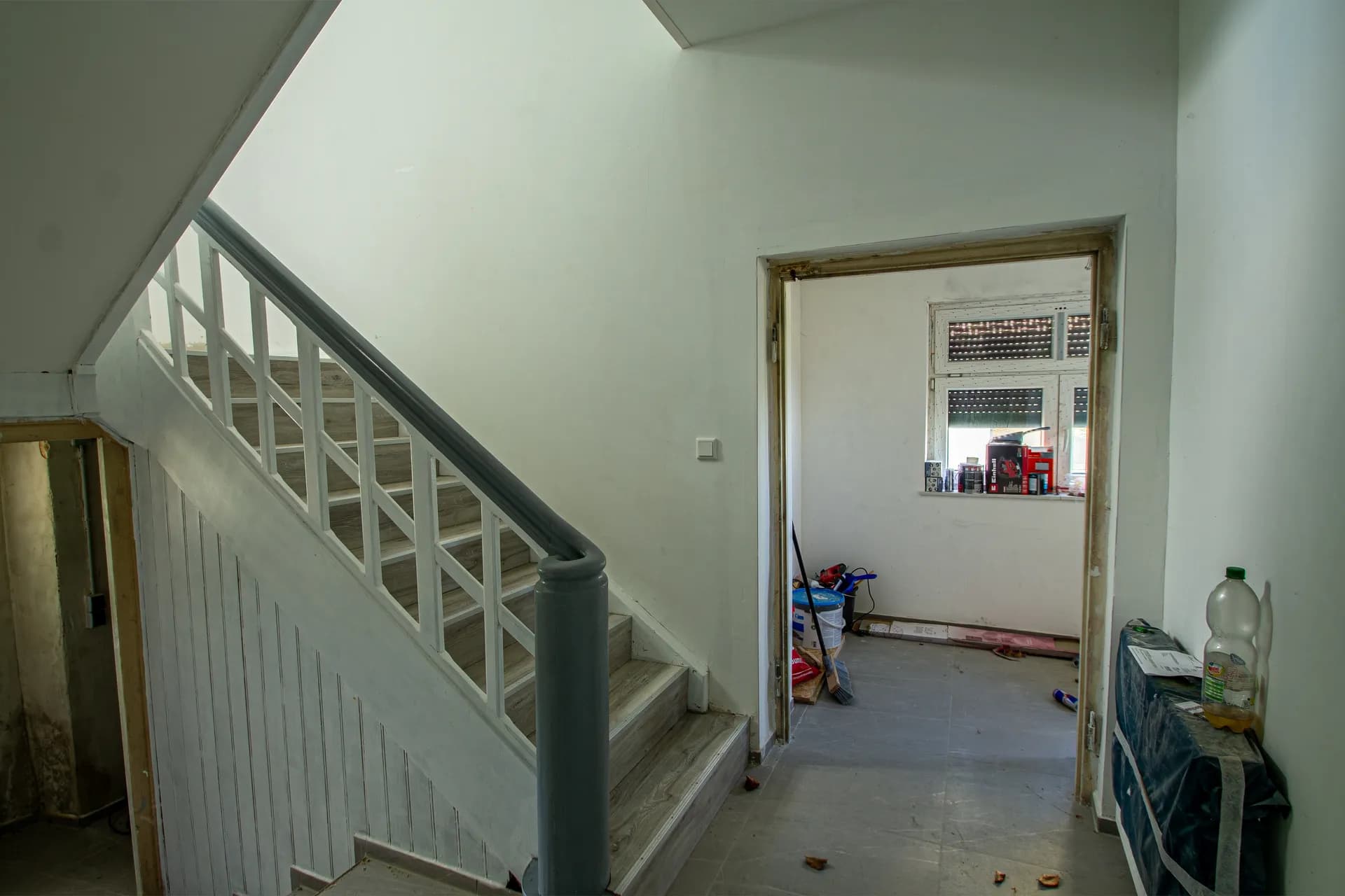 Eingangsbereich und Treppenaufgang mit grau gestrichenem Geländer und Durchgang zu einem renovierungsbedürftigen Zimmer.