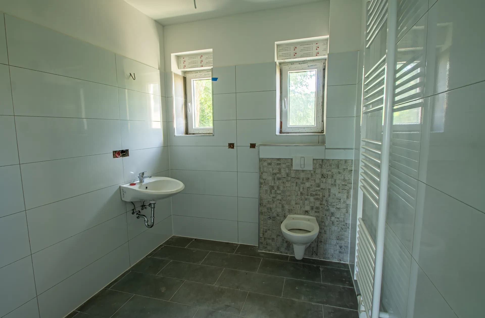 Helles Badezimmer mit dunklen Bodenfliesen, weißen Wandfliesen, Hänge-WC, Waschbecken und zwei Fenstern.