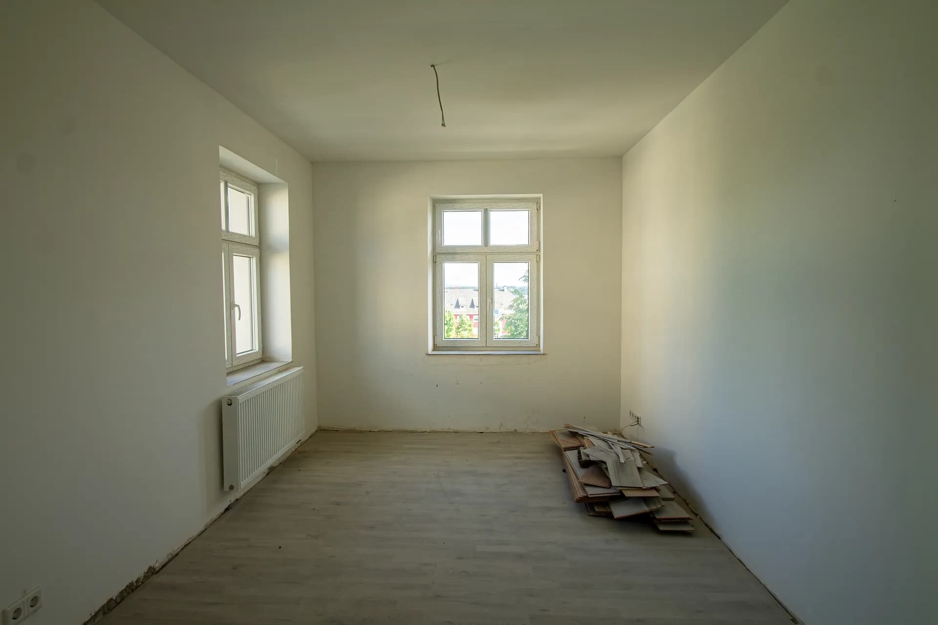 Lichtdurchfluteter Raum während der Renovierung mit zwei Fenstern über Eck, Heizkörper und restlichen Baumaterialien auf dem Boden.