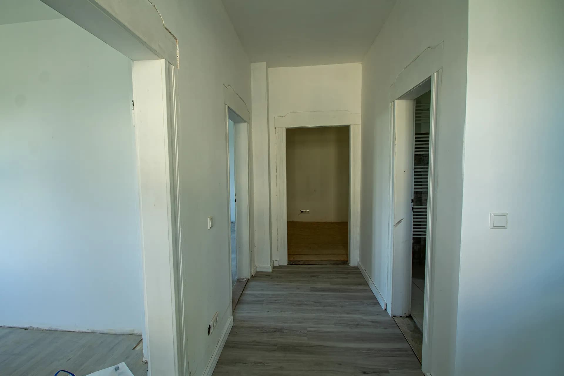 Wohnungsflur mit grauem Laminatboden, weiß gestrichenen Wänden und offenen Türdurchgängen zu den anliegenden Zimmern.