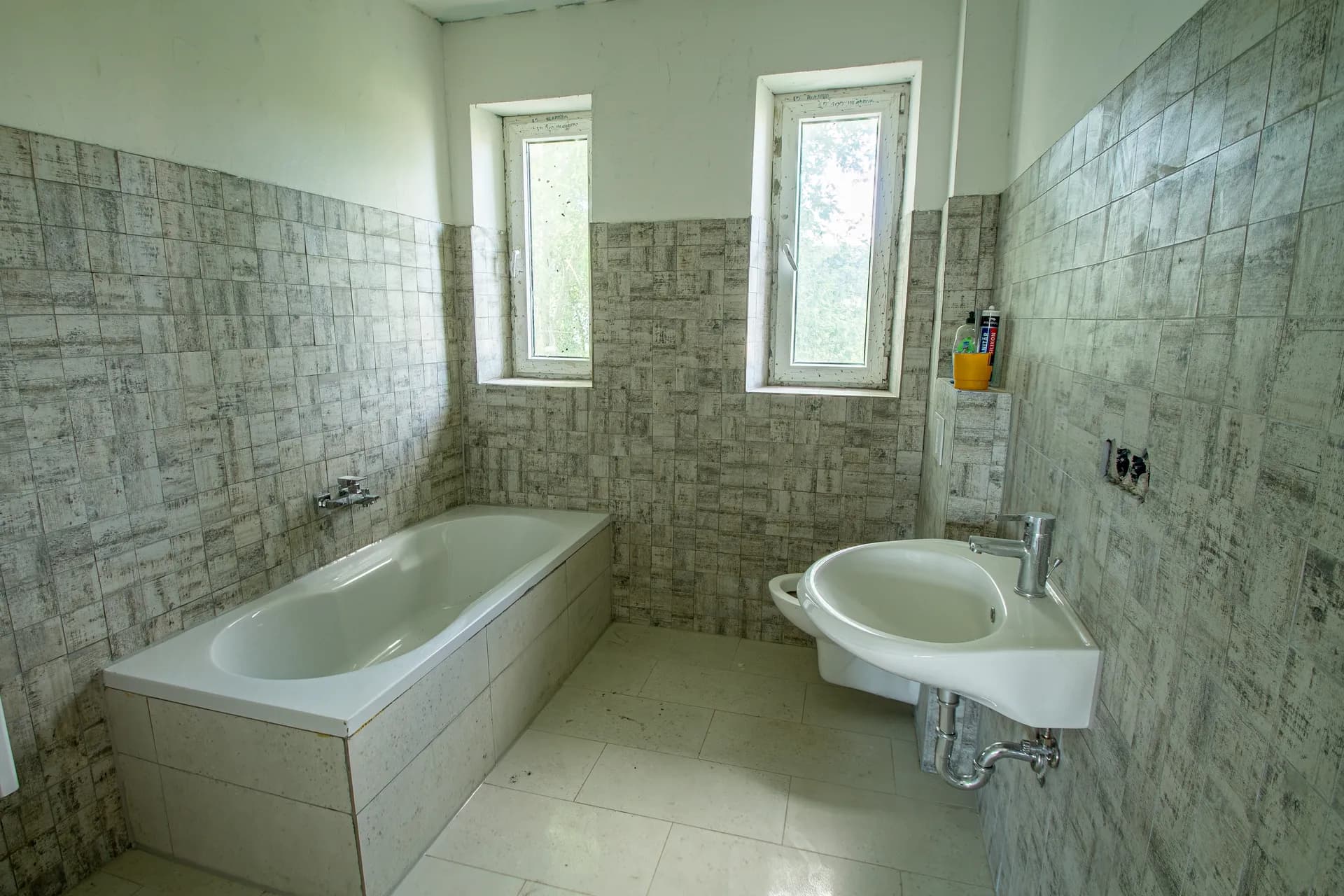Tageslichtbadezimmer mit grauer Struktur-Fliesung, Einbaubadewanne, Waschbecken und zwei Fenstern.