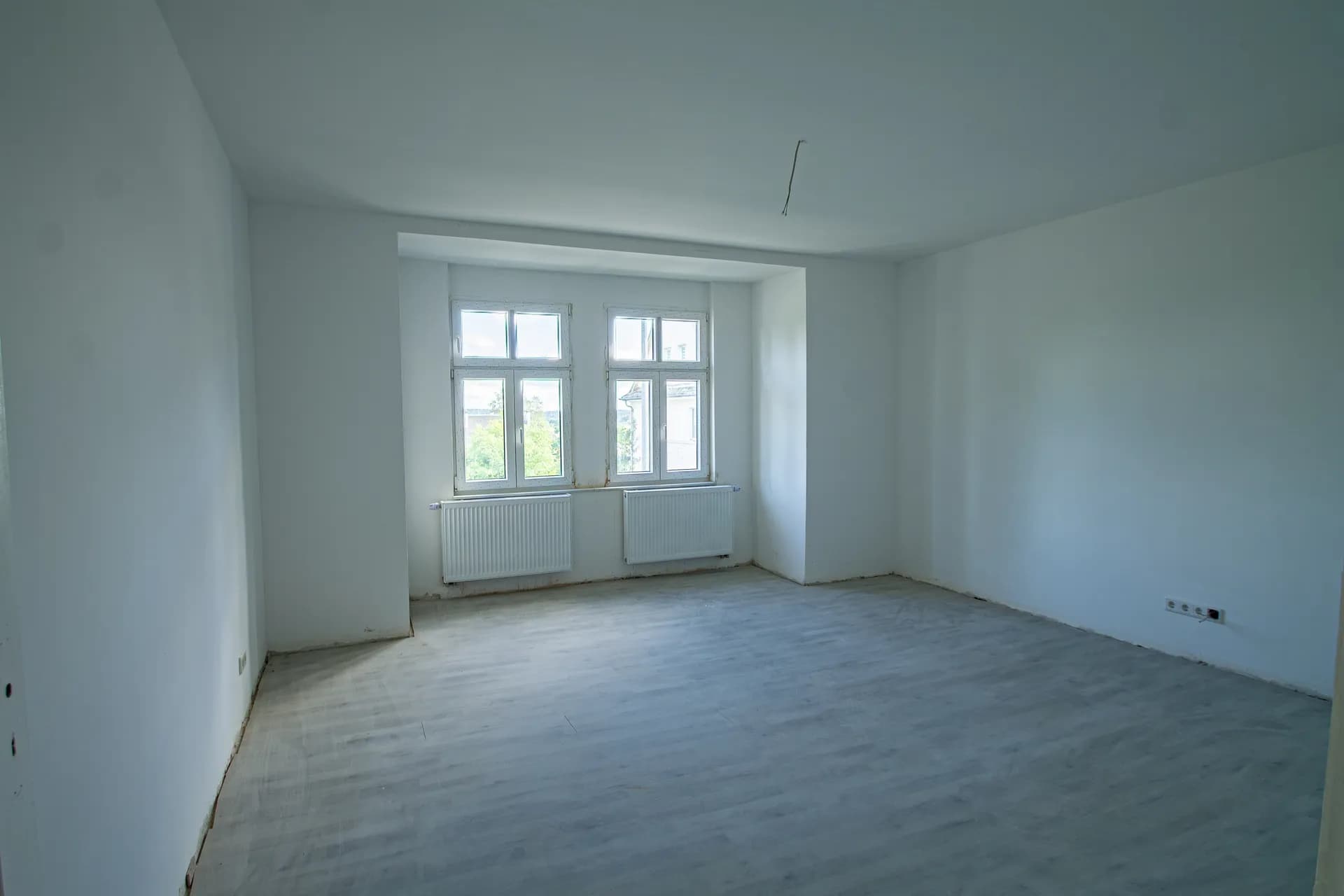 Großzügiges, lichtdurchflutetes Zimmer mit Erkerbereich, weiß verputzten Wänden, zwei Fenstern mit Heizkörpern und grauem Bodenbelag.