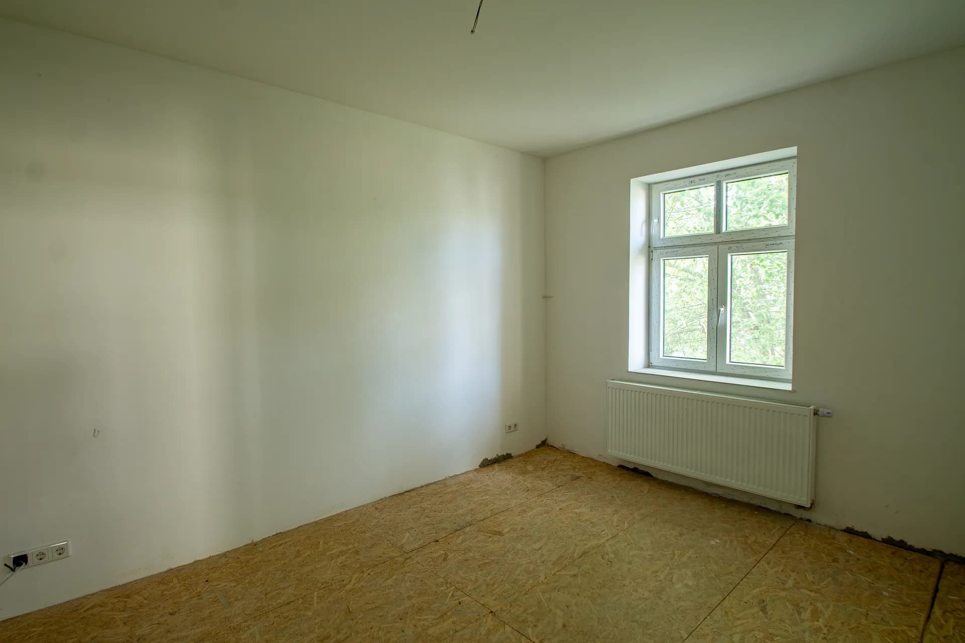 Helles, renovierungsbedürftiges Zimmer mit weiß verputzten Wänden, OSB-Verlegeplatten als Unterboden, Fenster und Heizkörper.