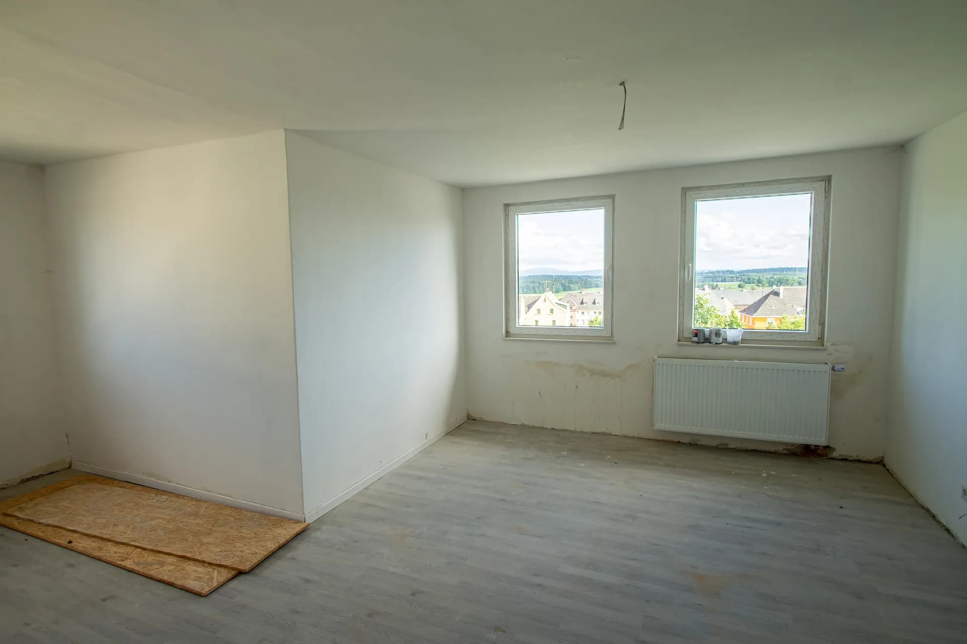 Heller, renovierungsbedürftiger Raum mit weißen Wänden, Laminatboden, zwei Fenstern mit Weitblick und Heizkörper.