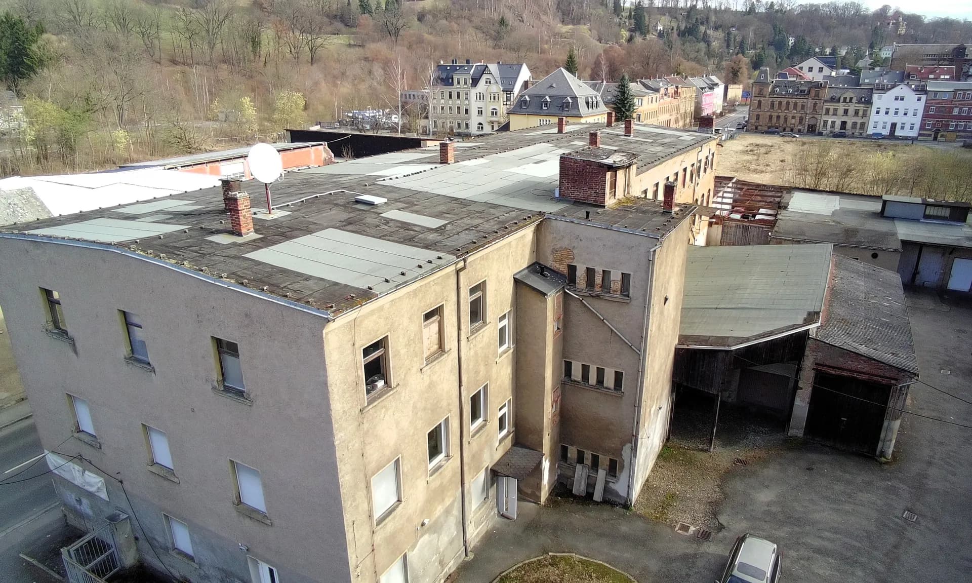 Luftaufnahme der rückwärtigen Ansicht des gewerblichen Gebäudekomplexes mit Flachdach, Anbauten, Lagerhallen und Blick auf die umliegende Tallage mit Stadtbebauung.