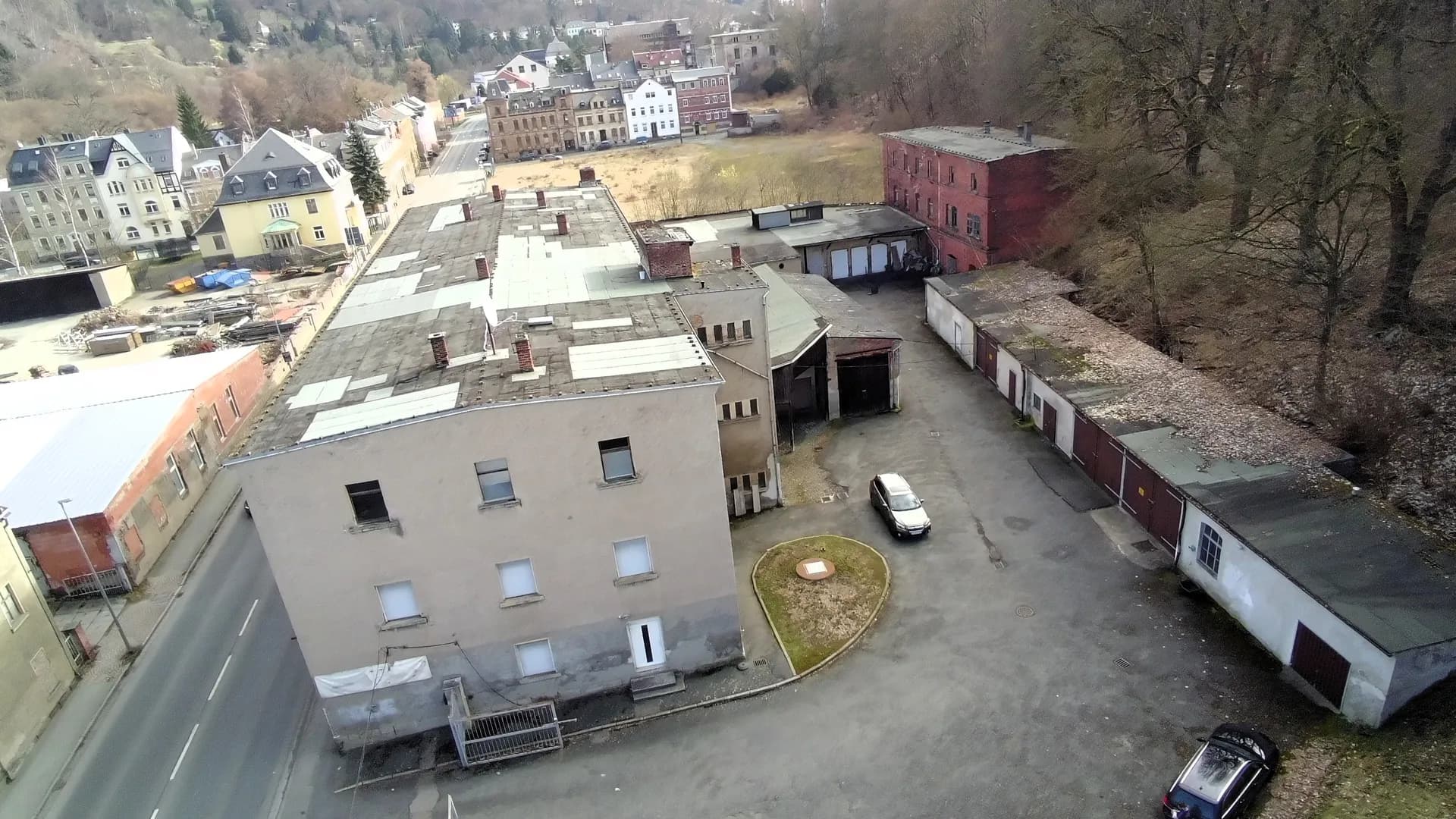 Übersichtliche Schrägluftaufnahme des gesamten Gewerbeareals mit Hauptgebäude, asphaltiertem Innenhof, Garagenzeile, historischem Ziegelbau und bewaldetem Hang im Hintergrund.