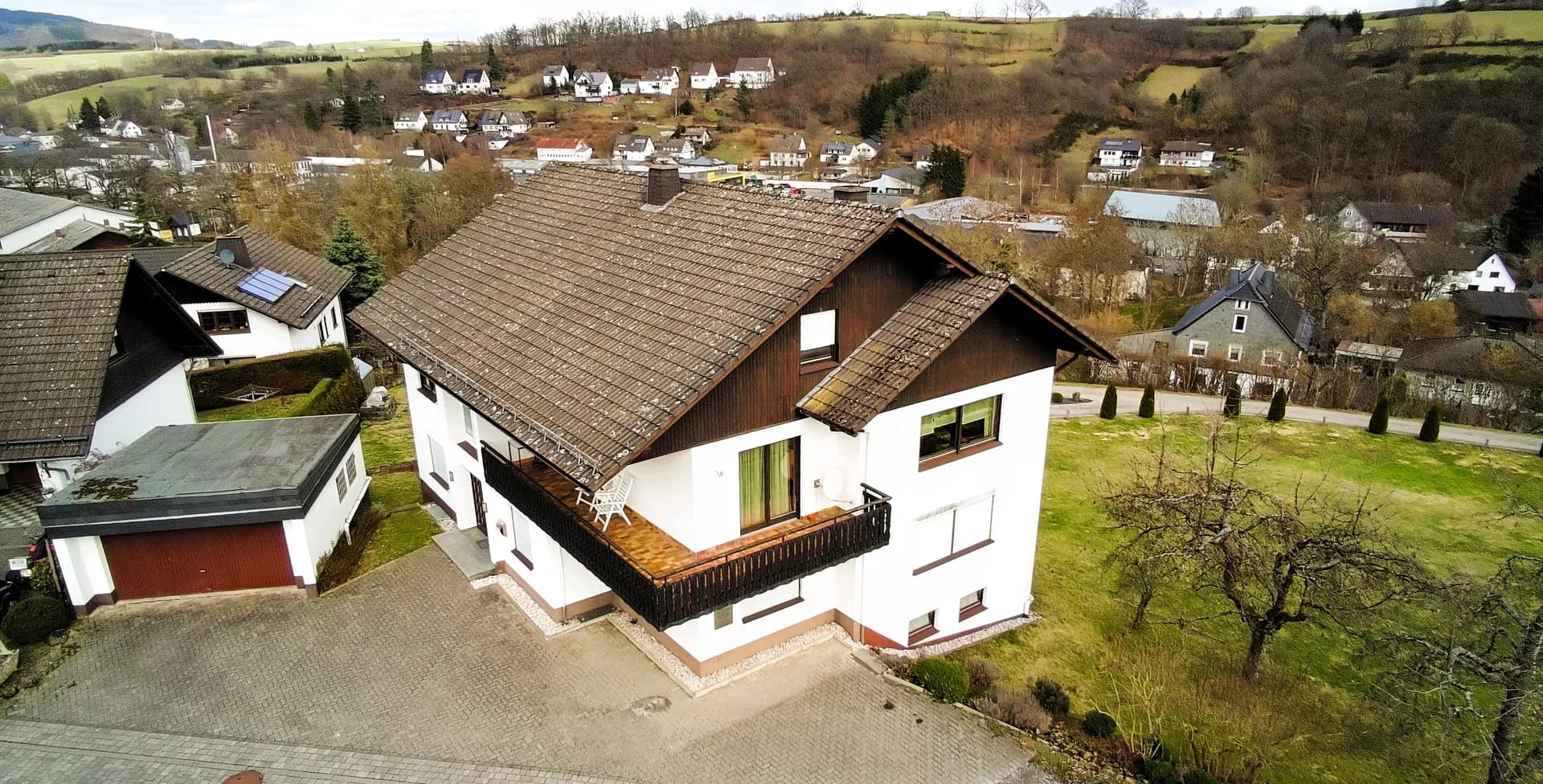 Luftbild-Vorderansicht des Wohngebäudes mit überdachtem Eingang, umlaufendem Balkon mit Sitzgelegenheiten, Garagentrakt und gepflasterter Hofeinfahrt.