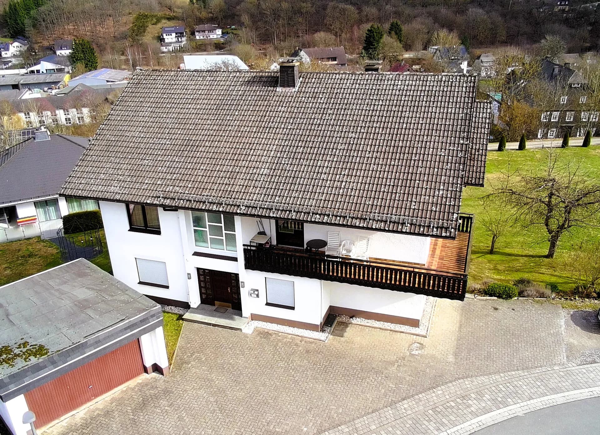 Luftbild-Vorderansicht des Wohngebäudes mit überdachtem Eingang, umlaufendem Balkon mit Sitzgelegenheiten, Garagentrakt und gepflasterter Hofeinfahrt.