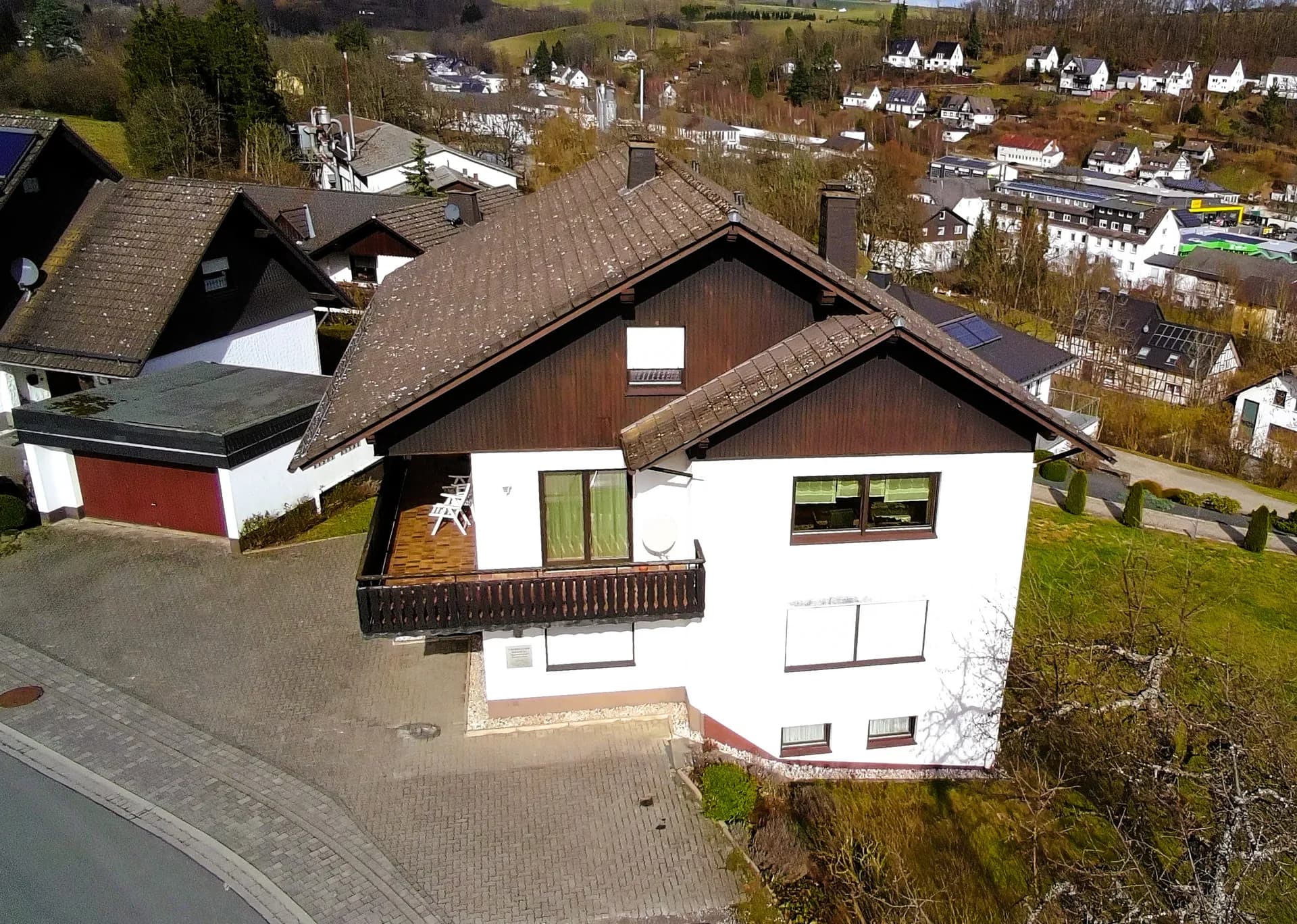 Schrägluftaufnahme des gepflegten Einfamilienhauses mit Giebel-Holzverschalung, großem Balkon, gepflastertem Hofraum, Garage und malerischem Ausblick über die Tallage.